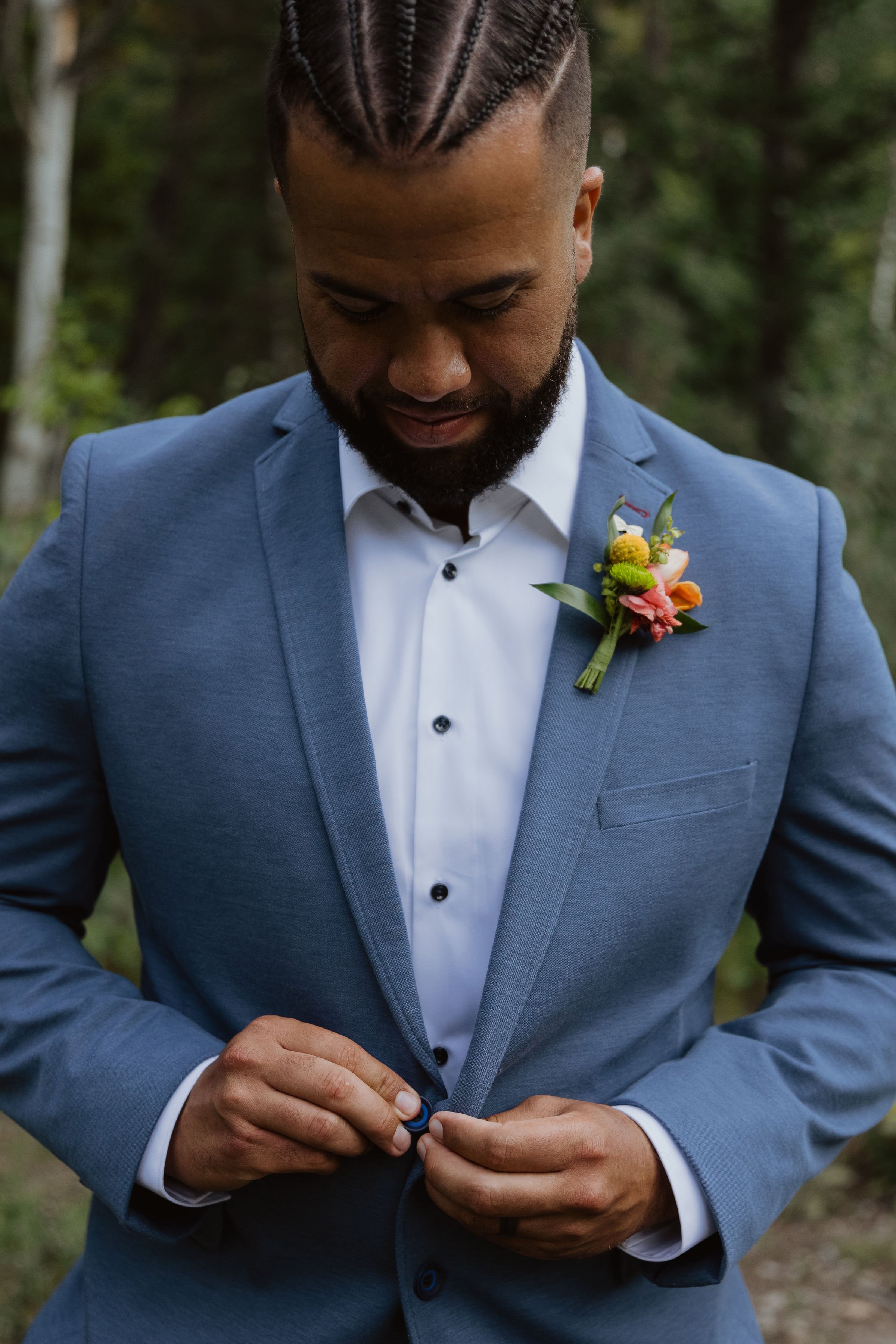 A man in a blue suit and white shirt is buttoning his suit jacket with a boutonniere on the lapel. He has braided hair and a beard, standing outdoors with green trees in the background.