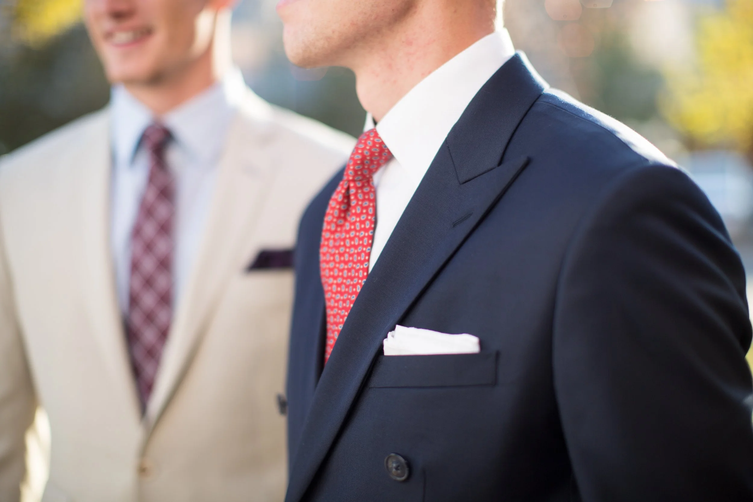 Close-up of two men dressed in formal business suits, one in a navy suit with a red tie and pocket square, the other in a cream suit with a patterned tie, both smiling outdoors on a sunny day.