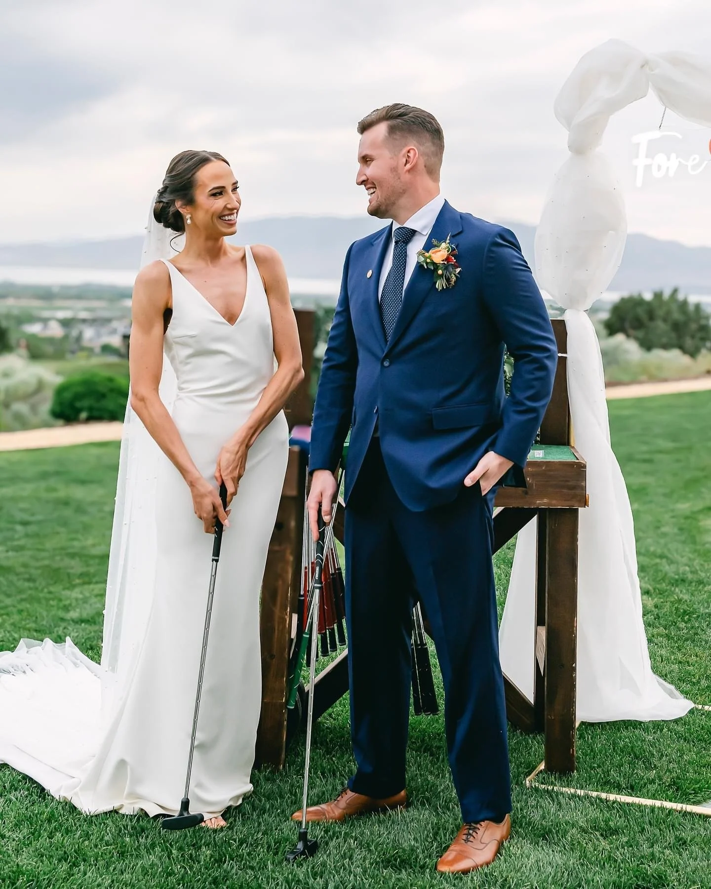 A bride and groom standing outdoors at a wedding ceremony, smiling at each other, with golf clubs in front of them and a white draped arch behind them.