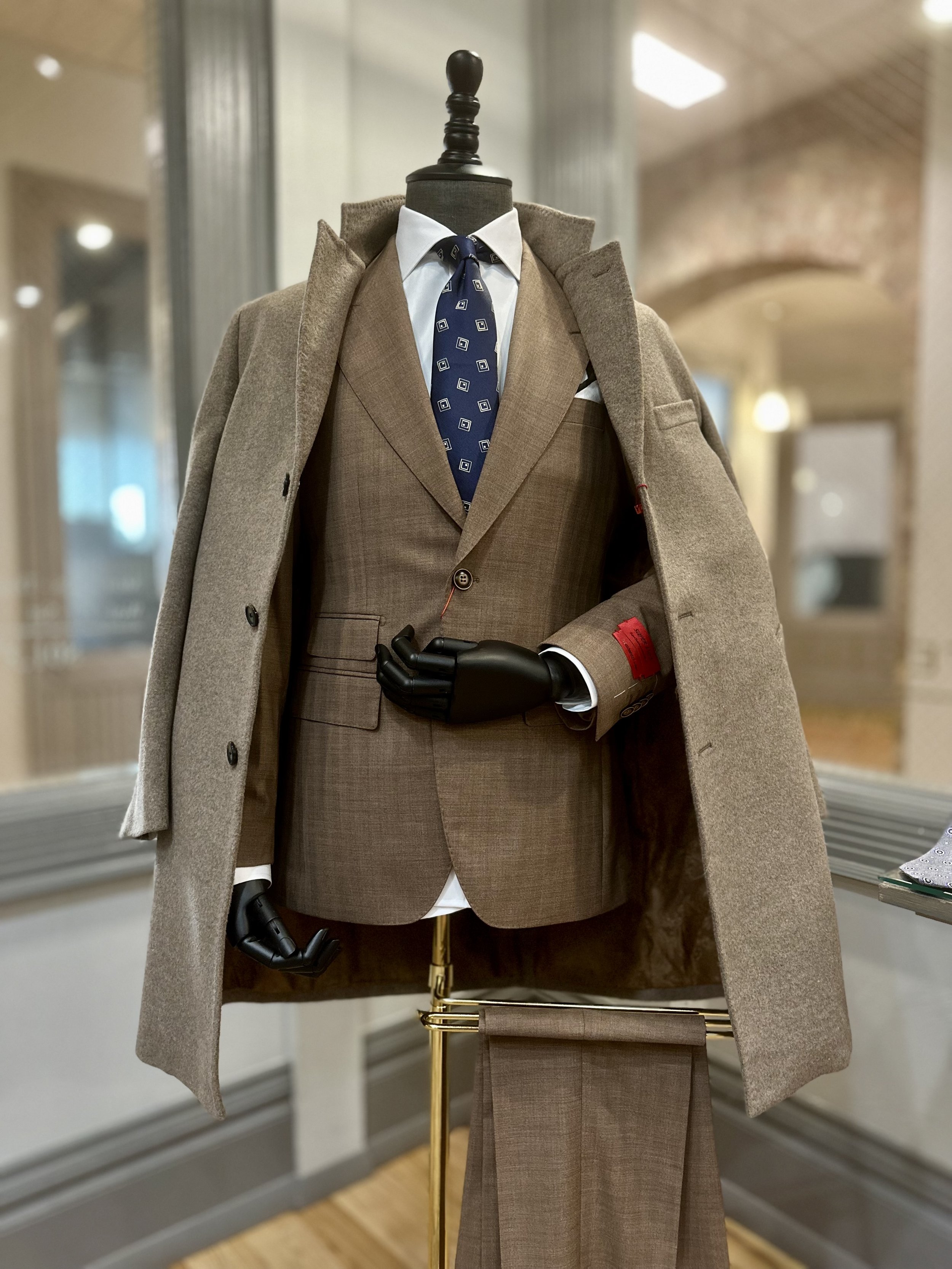 A mannequin dressed in a brown suit, white shirt, and navy patterned tie, with a camel overcoat draped over its shoulders, displayed in a retail store.