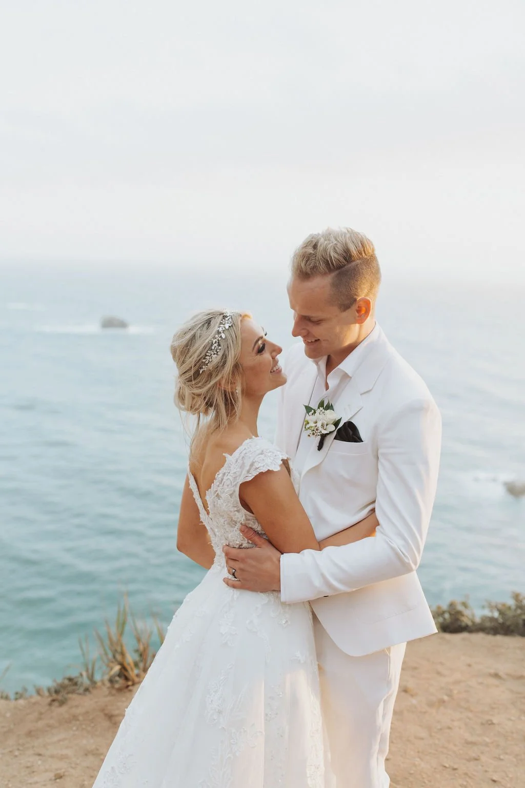 A bride and groom in wedding attire smiling and looking at each other by the ocean with a rocky coastline in the background.