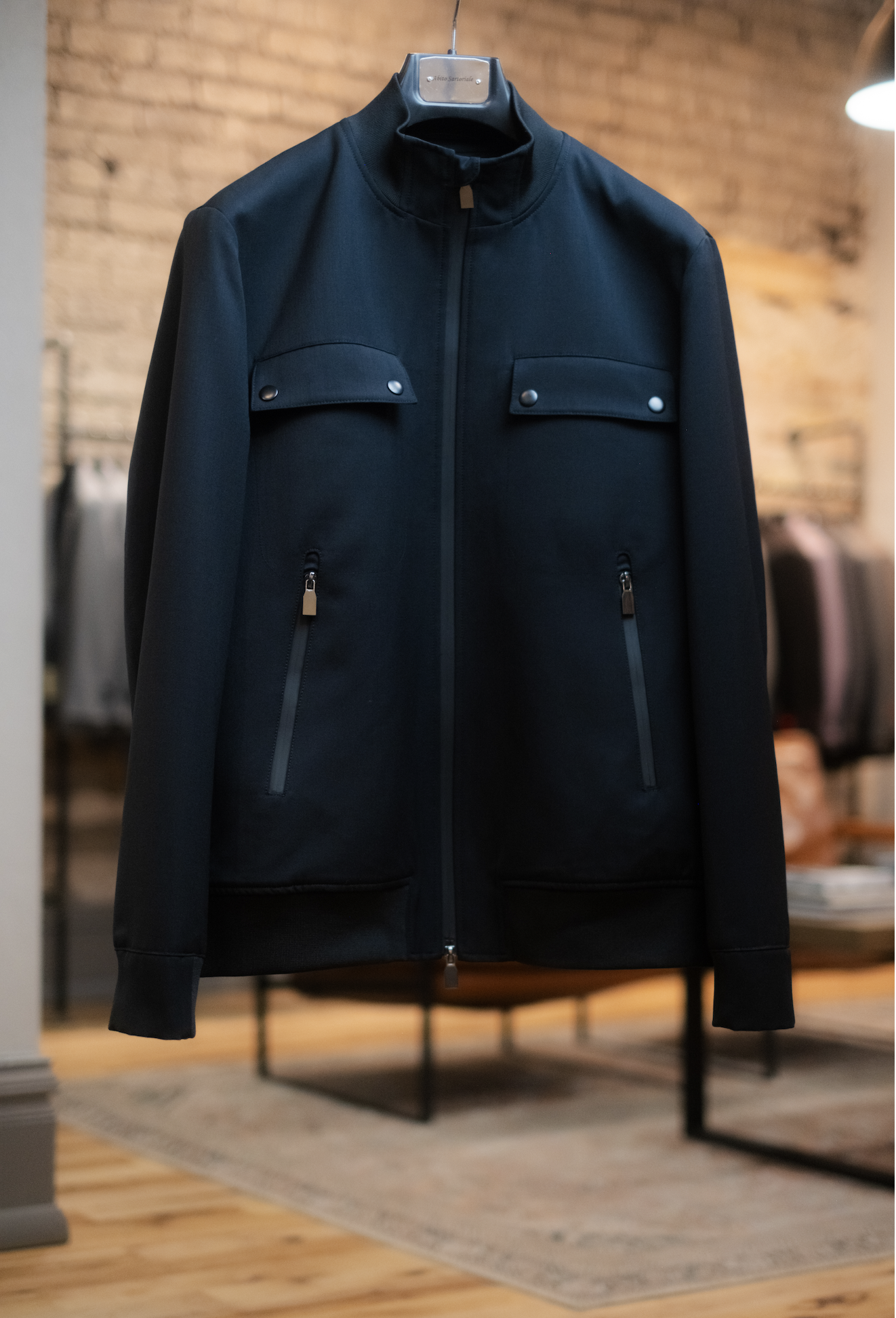 A black men's jacket with a standing collar, front zipper closure, and two zipper pockets, hanging on a hanger in a store with a brick wall background.