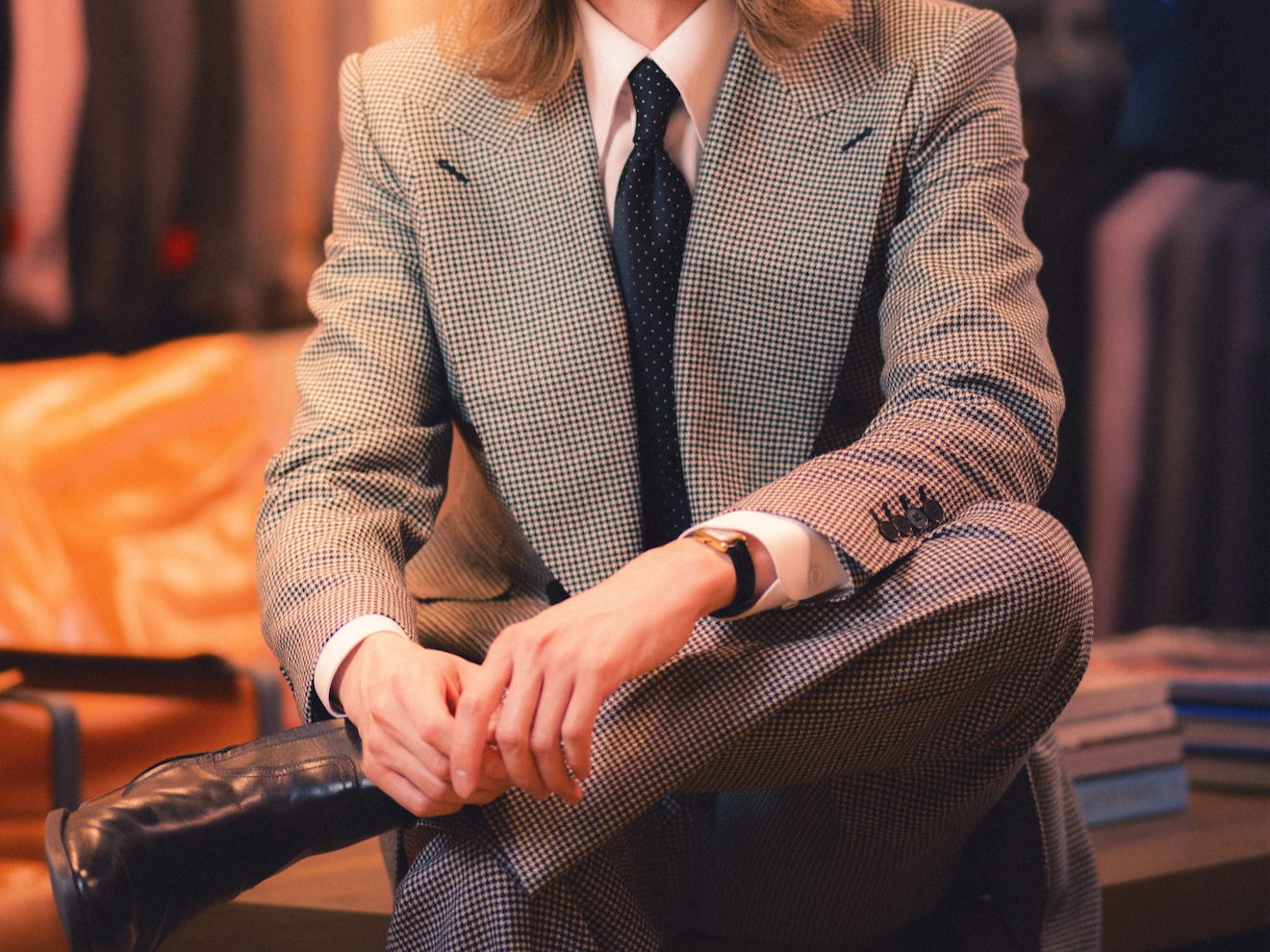 A person wearing a checkered suit with a tie, sitting with one leg crossed over the other, in a cozy, well-decorated room with books and warm lighting.