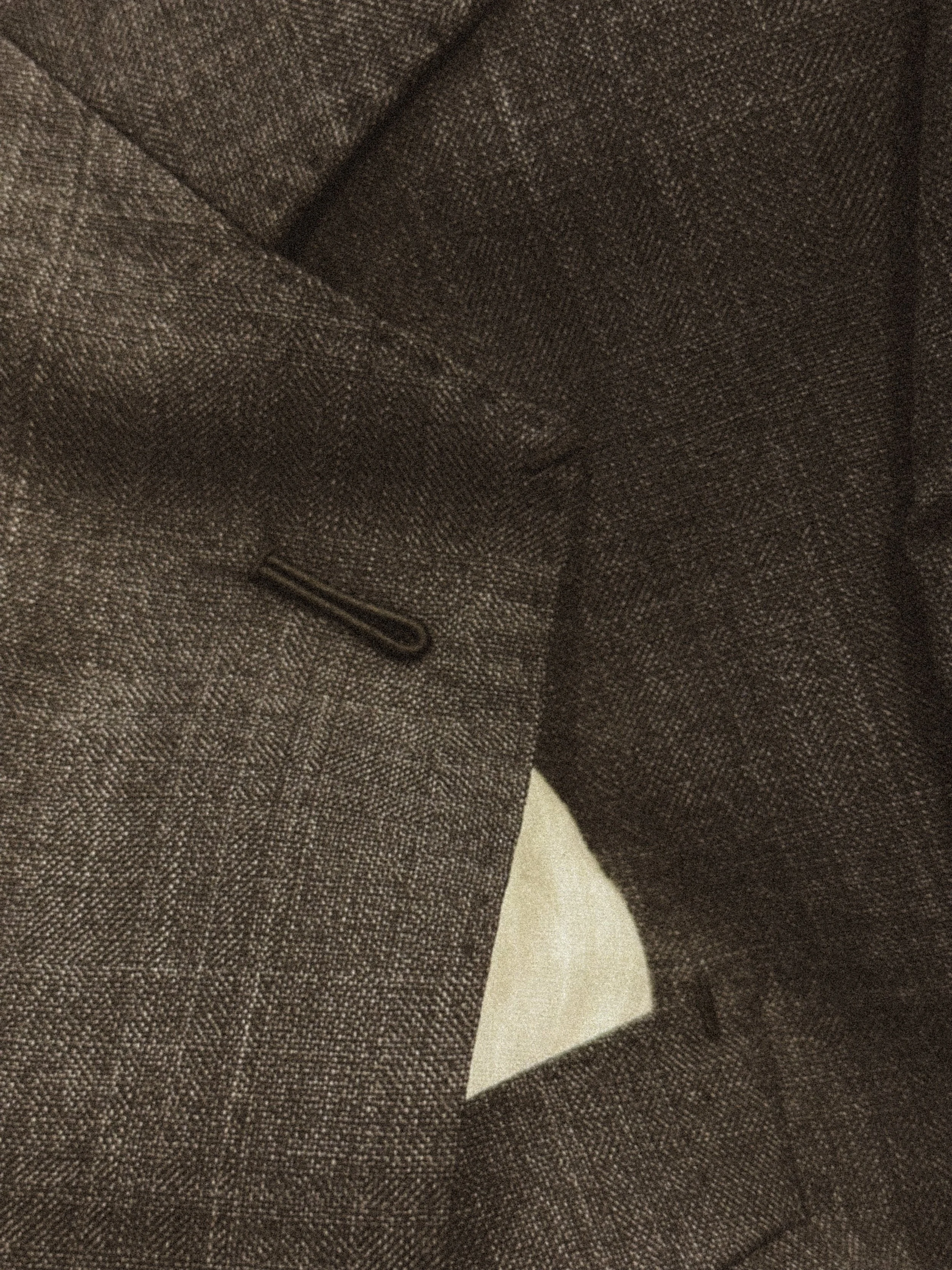 Close-up of charcoal colored tailored suit jacket with textured fabric, featuring a pocket and a stitching detail.
