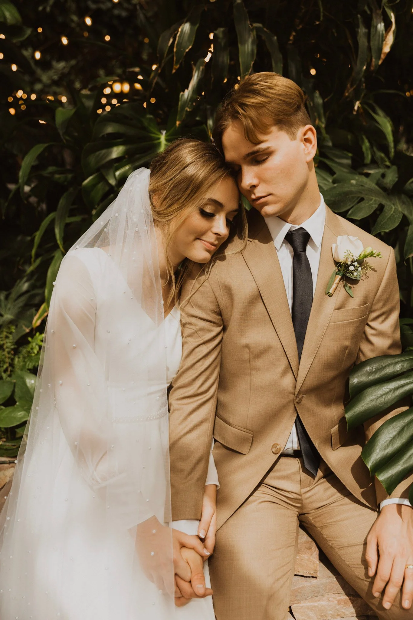 A bride and groom holding hands and with their foreheads touching in a tender moment during their wedding, standing in front of lush green foliage.