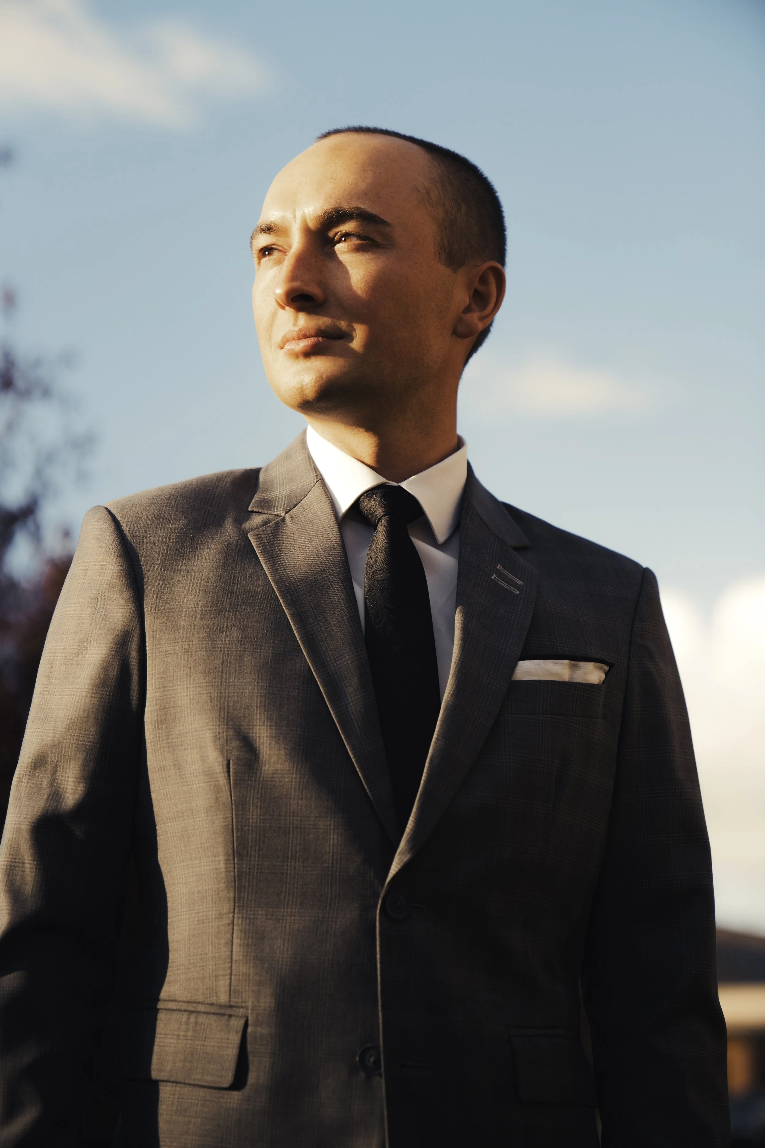 A man in a gray business suit and black tie standing outdoors during sunset, with a clear blue sky and some clouds in the background.