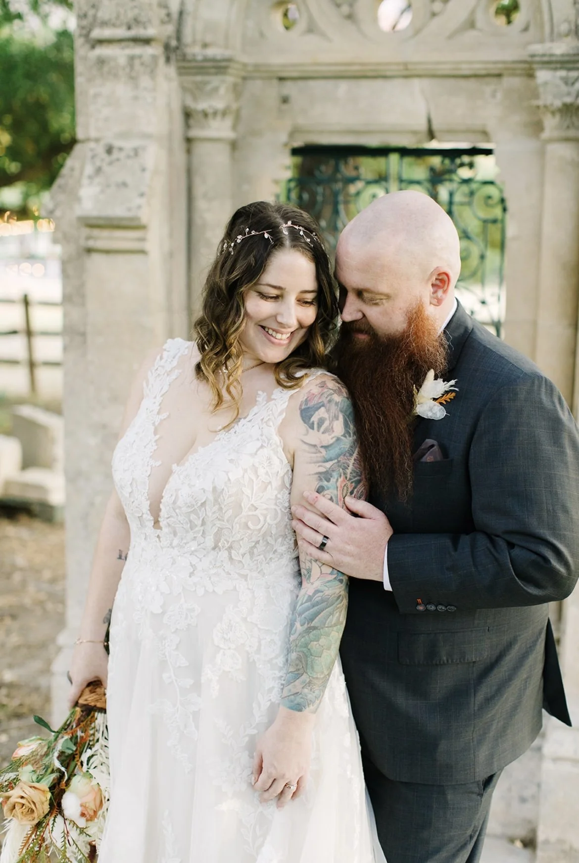 A happy woman in a white lace wedding dress and a man in a dark suit, standing close together outdoors near a stone gate, sharing a tender moment with the woman smiling and the man gently touching her shoulder.