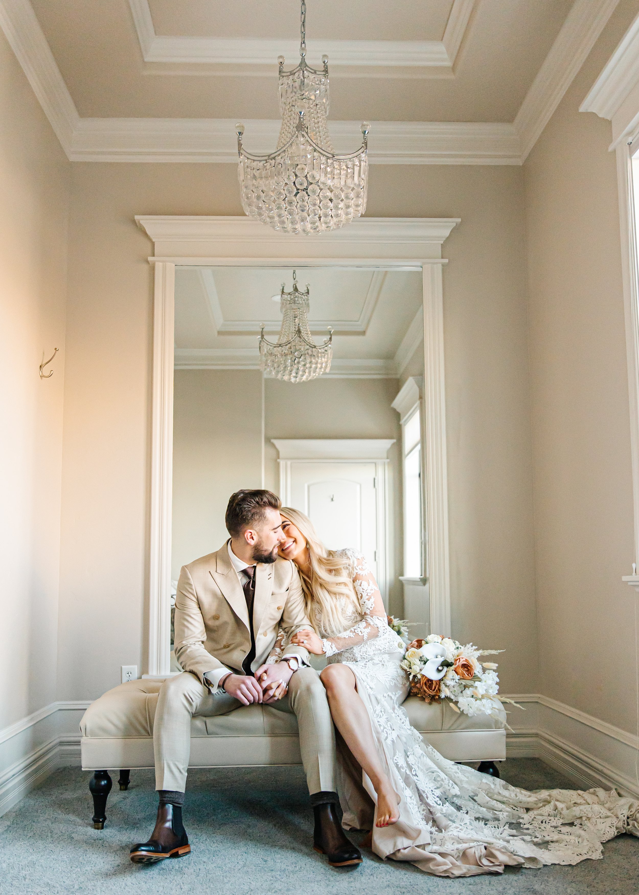 A bride and groom sitting on a bench in a room with a large mirror and chandelier, sharing a tender moment.