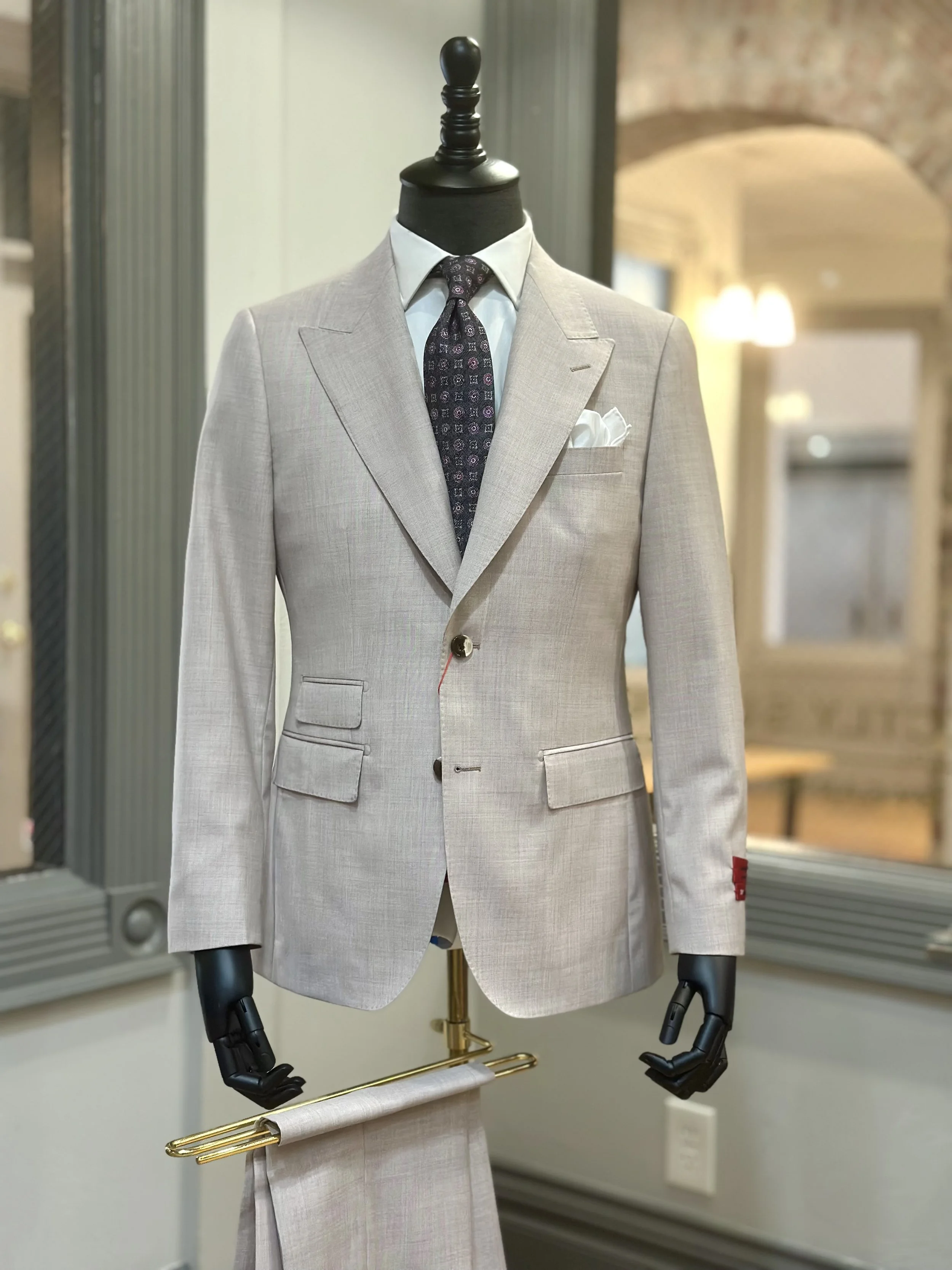 A mannequin dressed in a beige suit jacket with a white shirt, patterned tie, and pocket square, displayed in a store.