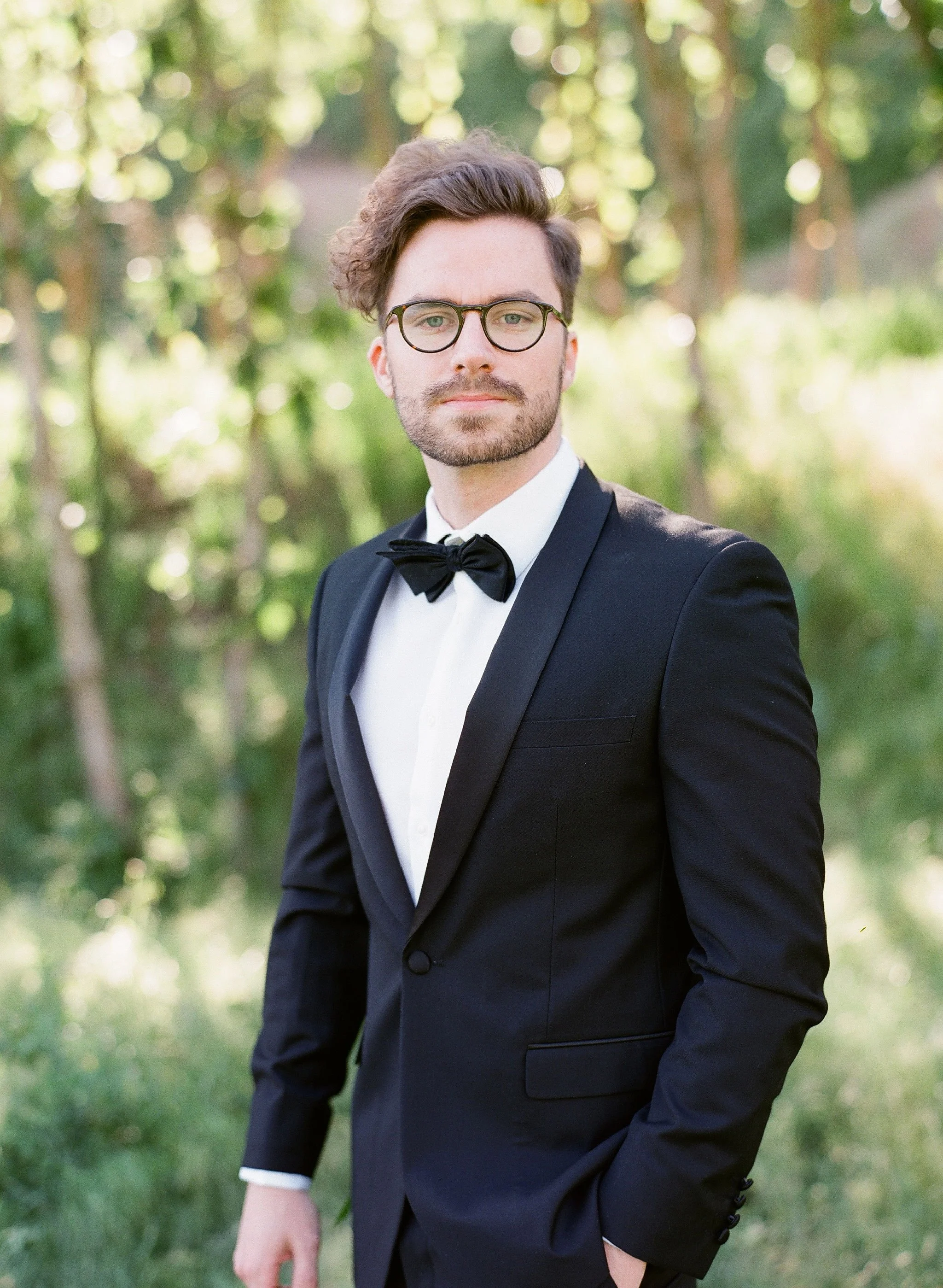 A man dressed in a black tuxedo with a white shirt and black bow tie, standing outdoors with a blurred green background, looking at the camera.