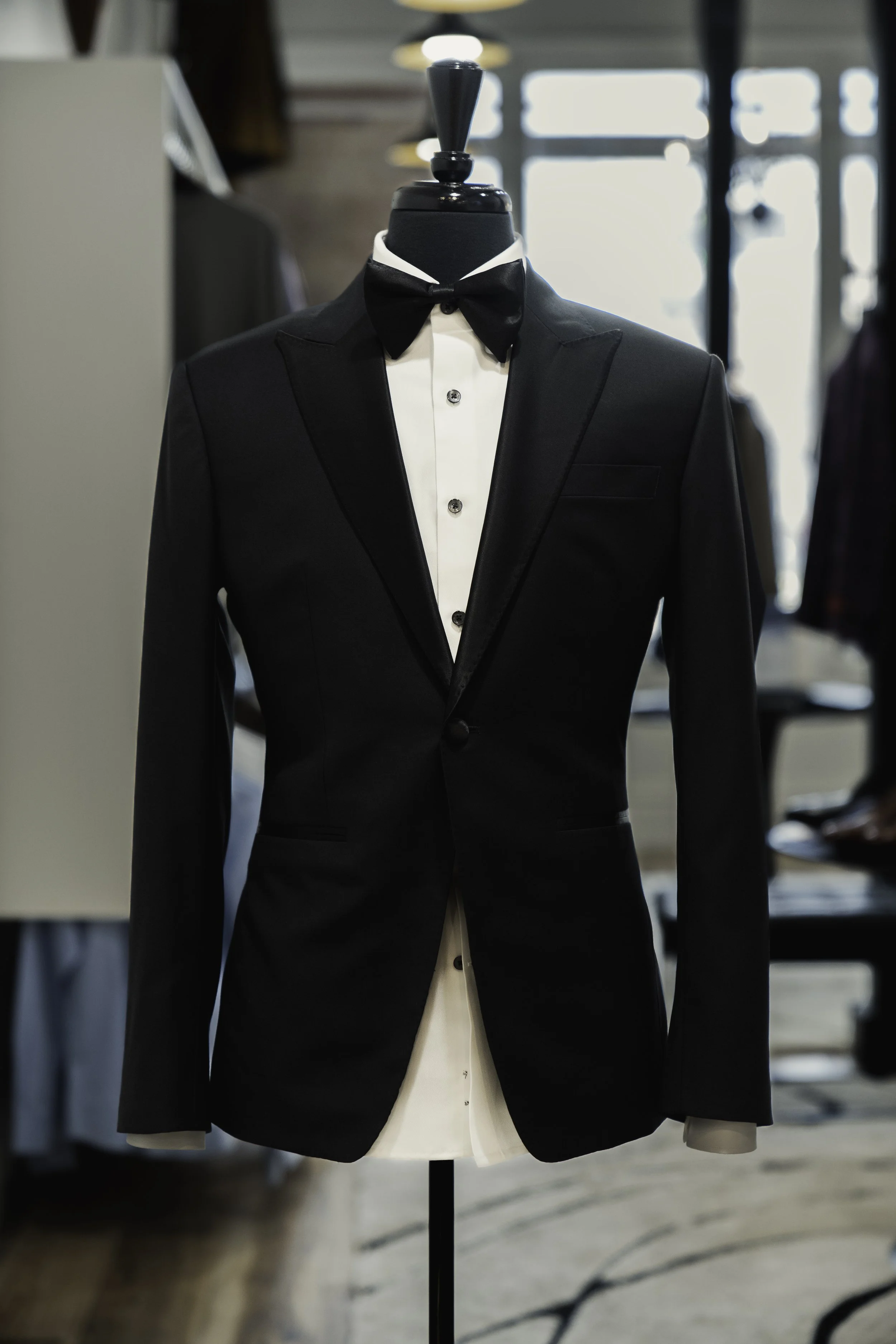 A tuxedo on a mannequin with a black jacket, white shirt, and black bowtie, inside a clothing store.