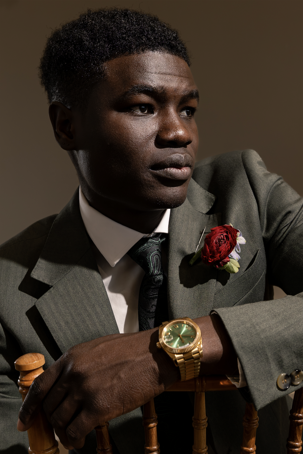 A young man in a green suit with a floral boutonniere, wearing a gold watch on his left wrist, is seated with his left arm resting on the back of a wooden chair. The background is neutral and the lighting highlights his face and attire.