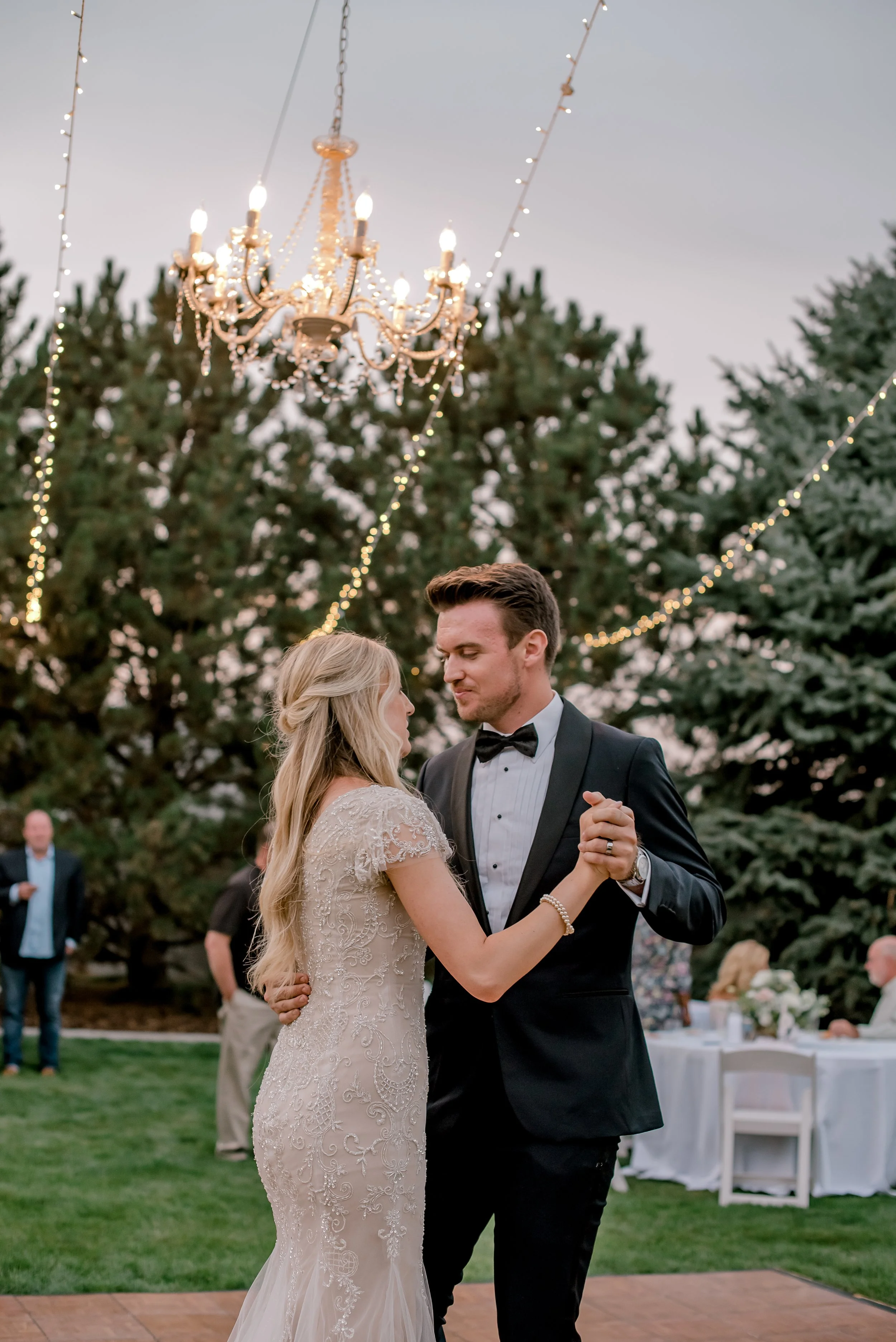 A bride and groom sharing a dance at an outdoor wedding reception, under string lights and a chandelier, with guests and greenery in the background.