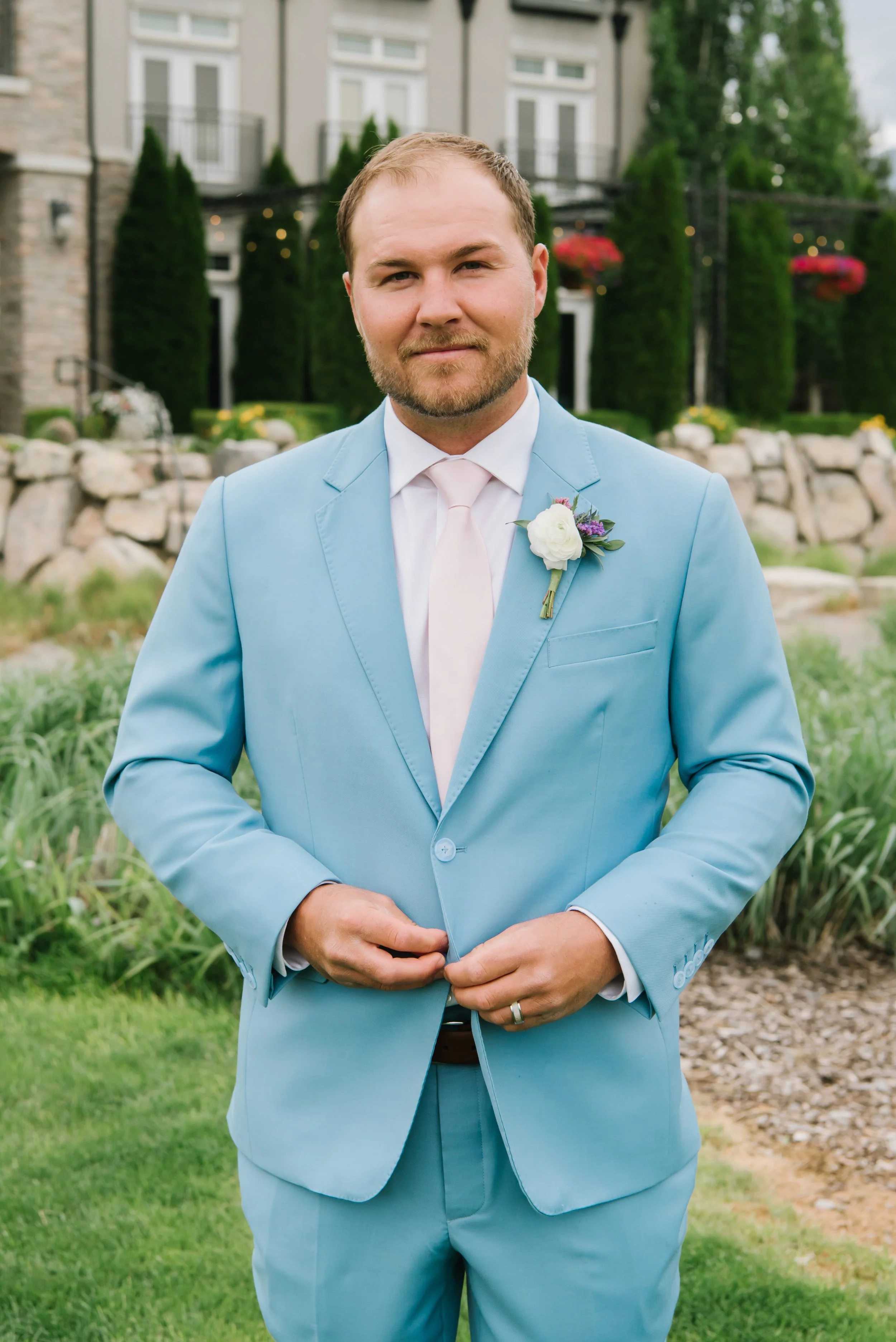 A man in a light blue suit with a white shirt and pink tie stands outdoors in front of a garden with green grass, rocks, and trees. He has a boutonniere on his lapel with a white flower and purple accents. The background features a building with balc