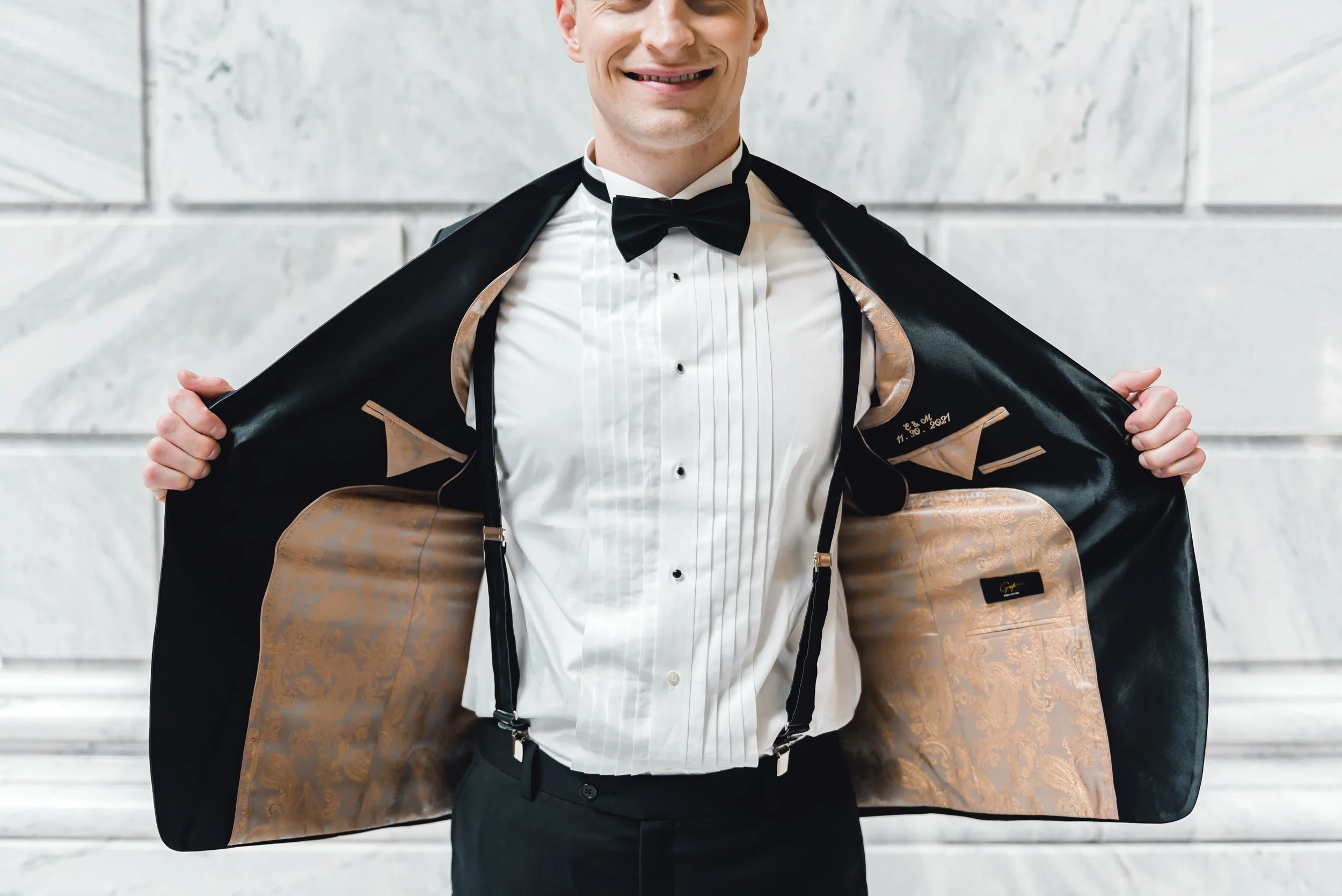 A man in a black tuxedo and bow tie is opening his tuxedo jacket to reveal a signature-lined, gold-embossed satin tuxedo lining, smiling in front of a marble wall.