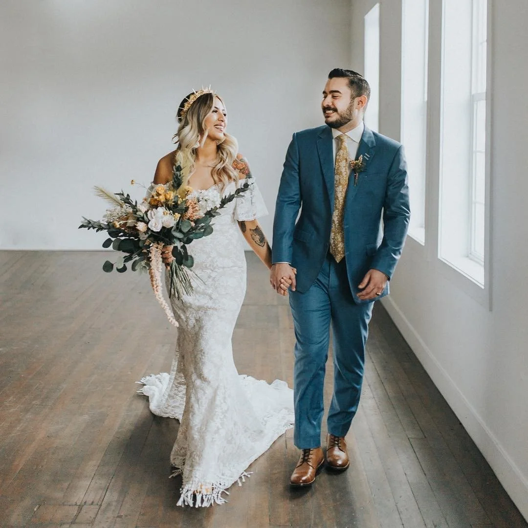 A bride and groom walking hand in hand, smiling, in a bright room with large windows. The bride is in a white lace wedding dress holding a bouquet of flowers, and the groom is in a blue suit with brown shoes.