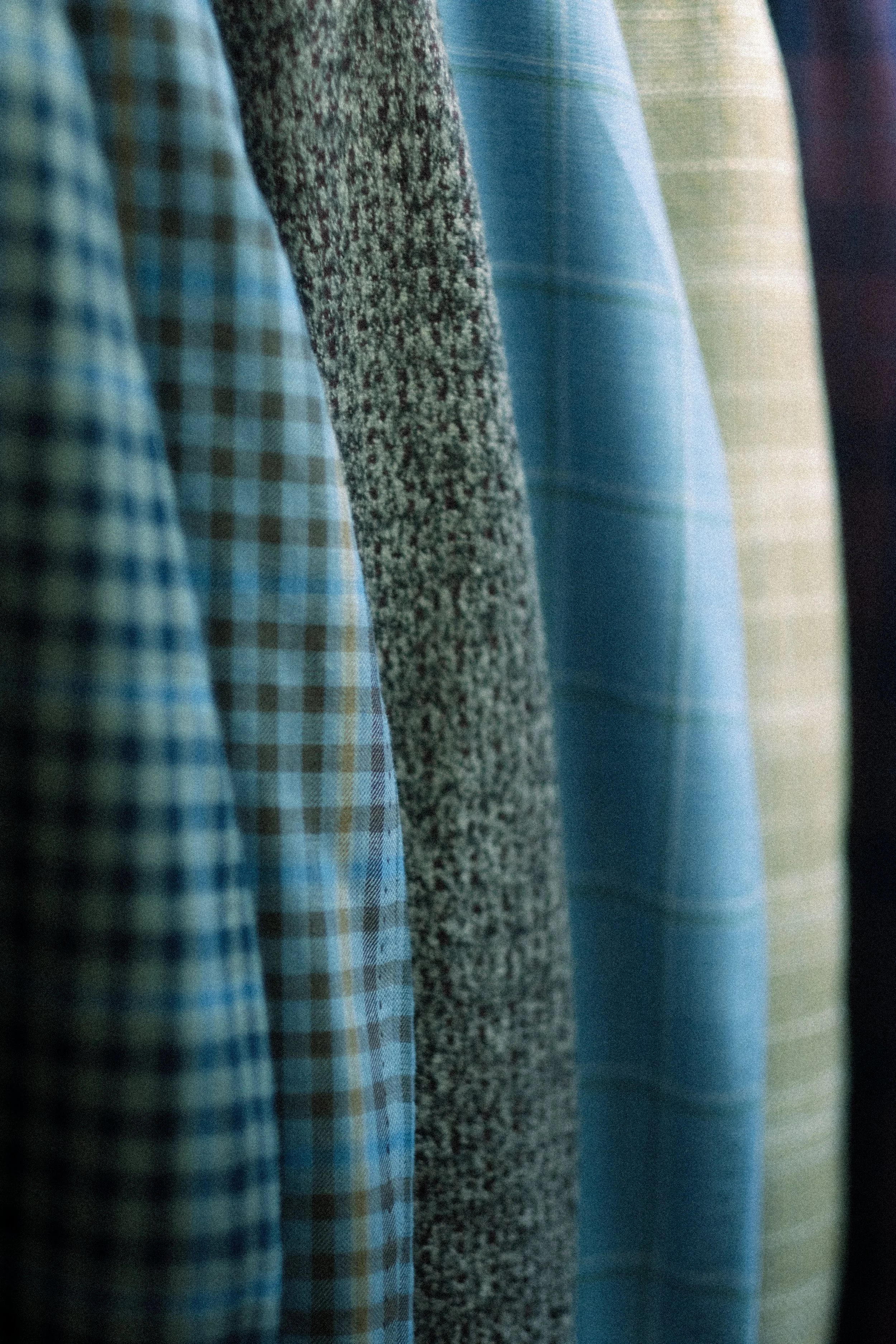 Close-up of various patterned and colored sport coats hanging on a rack, including plaid, and solid fabrics in shades of blue, green, and beige.