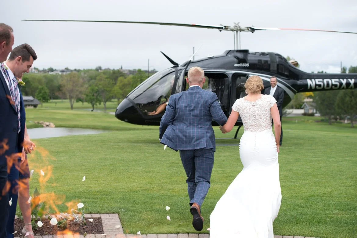 Bride and groom walking toward a helicopter, holding hands, with guests nearby during a wedding ceremony on a golf course.