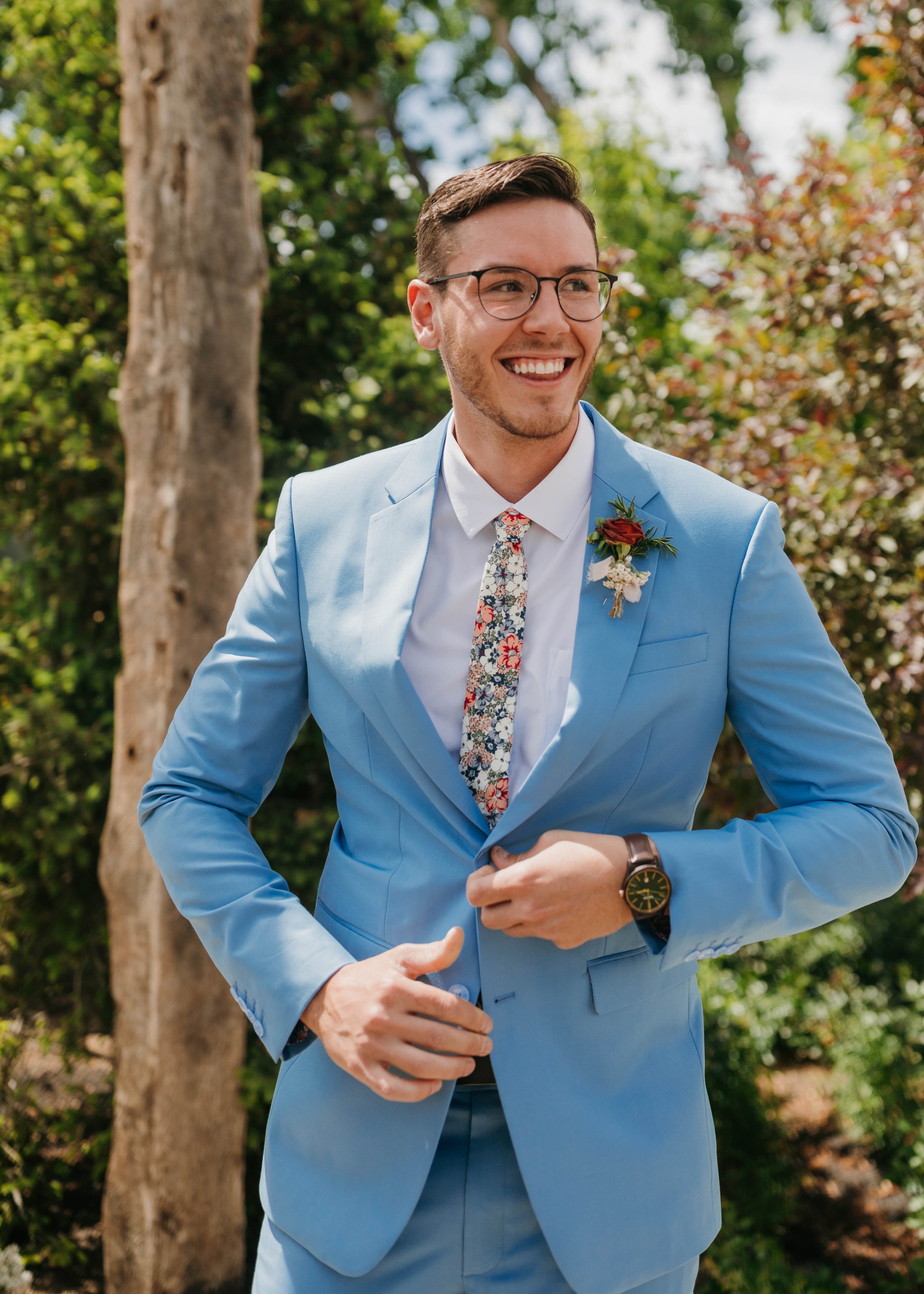 A young man in a light blue suit with a floral tie, glasses, and a boutonniere, smiling outdoors with trees and greenery in the background.