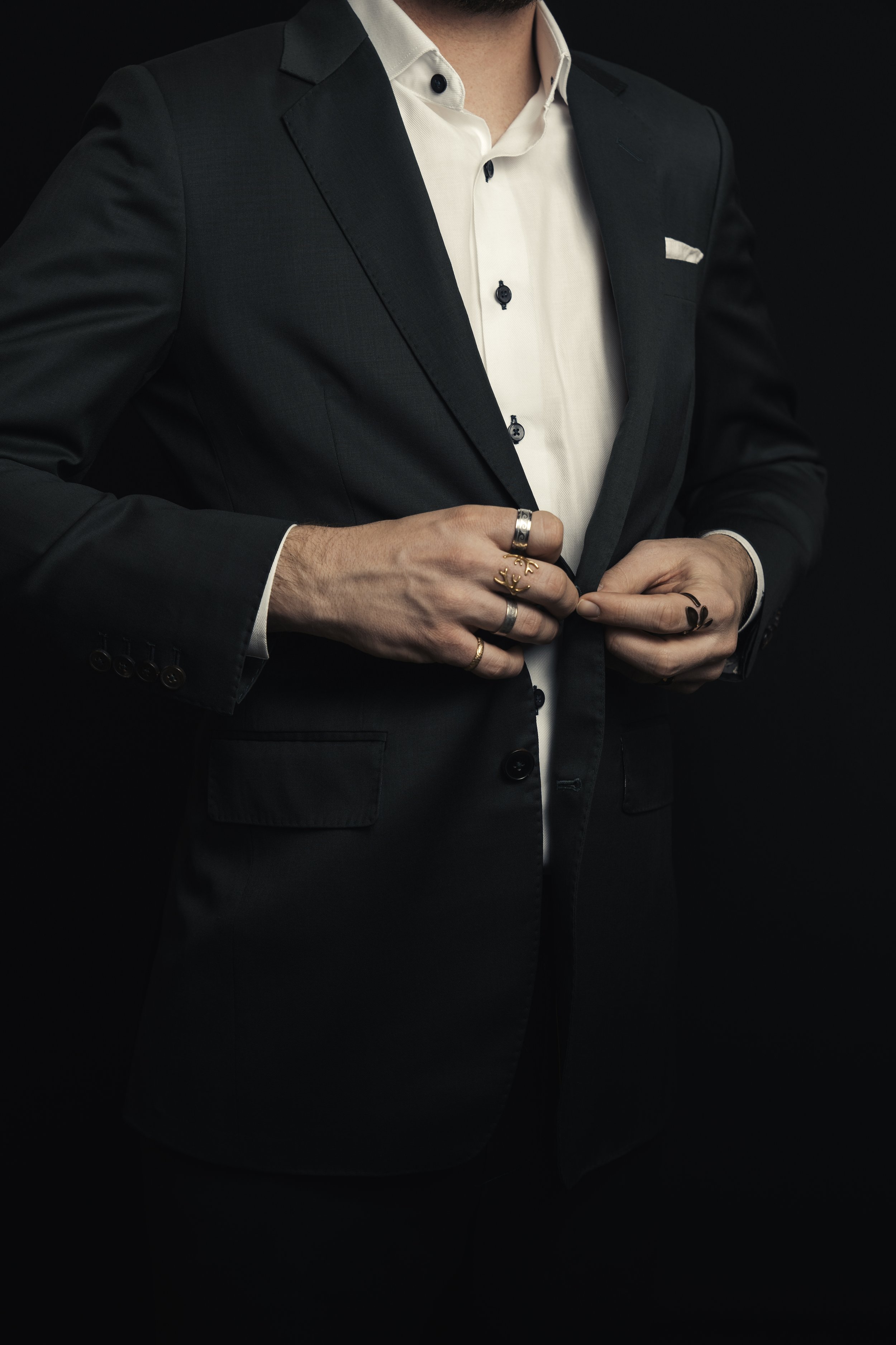 Close-up of a man wearing a dark green suit and a white dress shirt, with the focus on his torso and hands, against a black background.