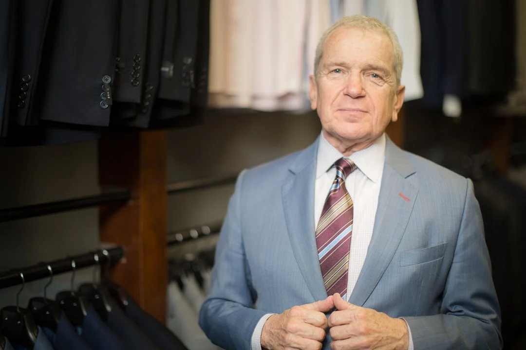 An elderly man in a light blue suit and tie standing in a clothing store with suits on hangers.