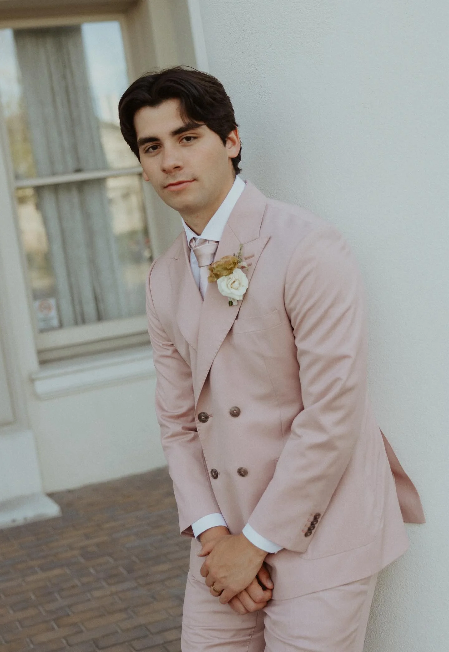 A young man with dark hair dressed in a light pink suit with a white shirt and boutonniere standing indoors near a white wall with windows behind him.
