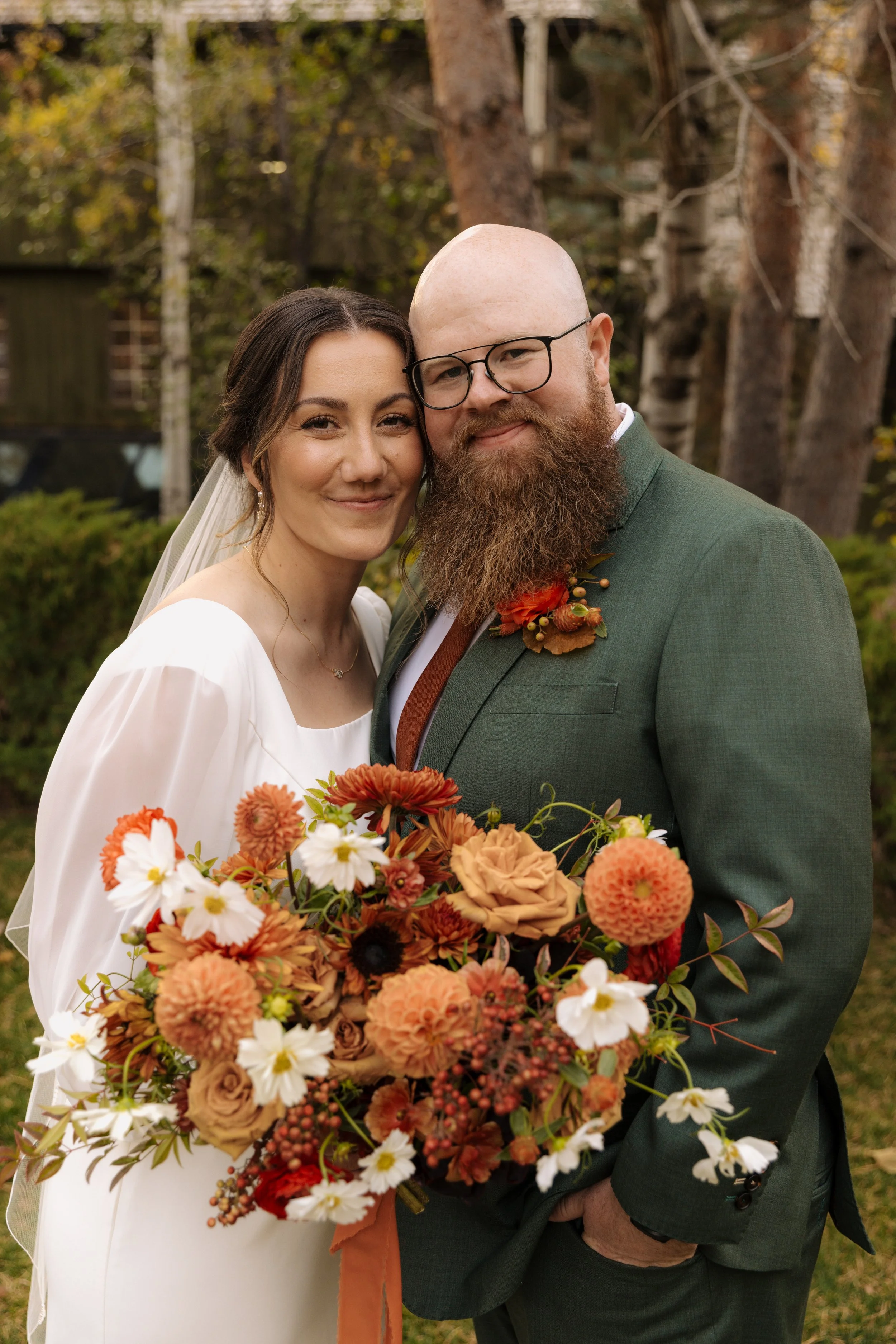 A bride and groom standing outdoors, smiling and close together. The bride is holding a large bouquet of orange, white, and brown flowers. The groom is wearing glasses and a green suit with a flower boutonniere. Trees and greenery are in the backgrou