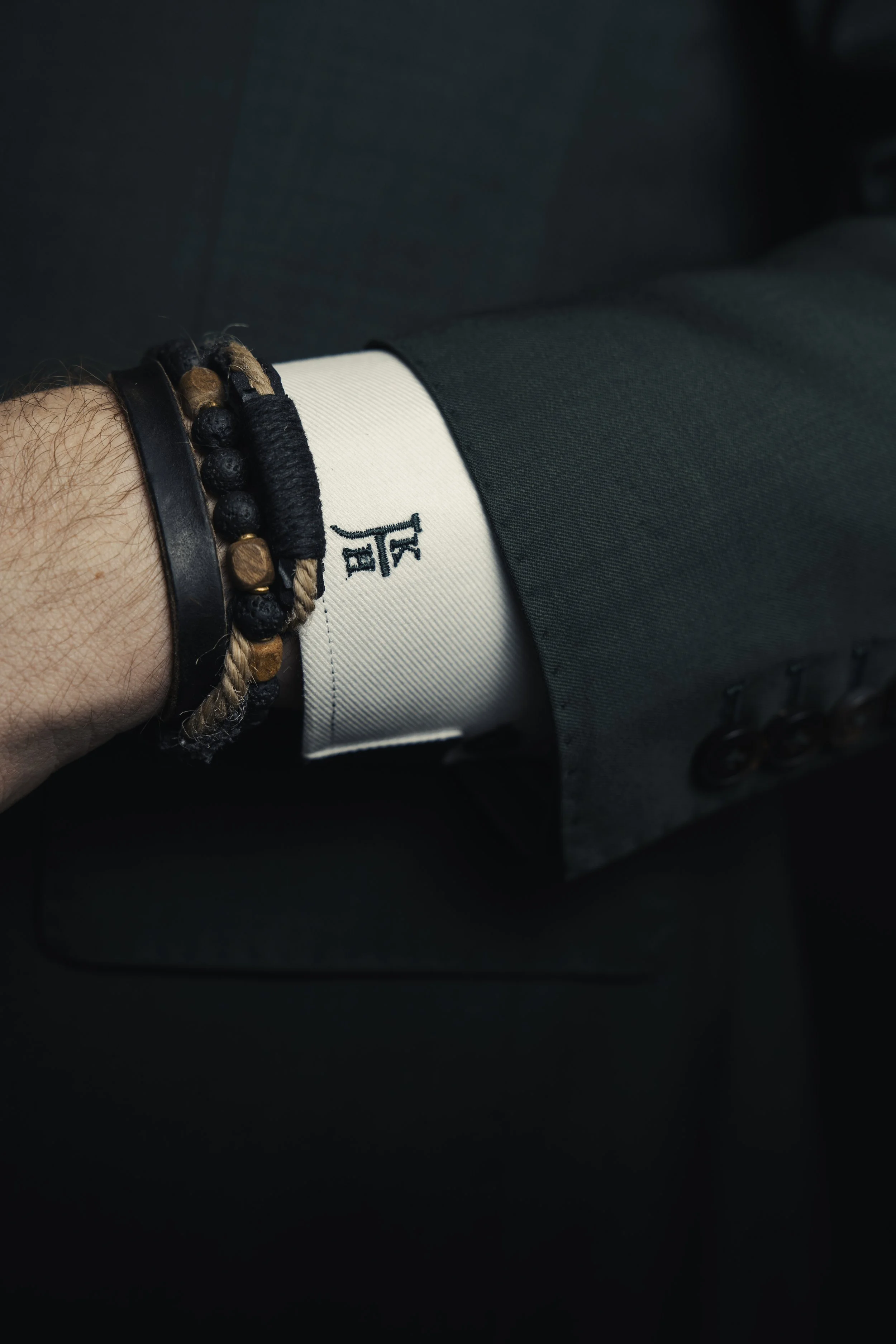 Close-up of a person's arm dressed in a suit, with a white shirt cuff, wearing multiple bracelets with beads and leather straps.
