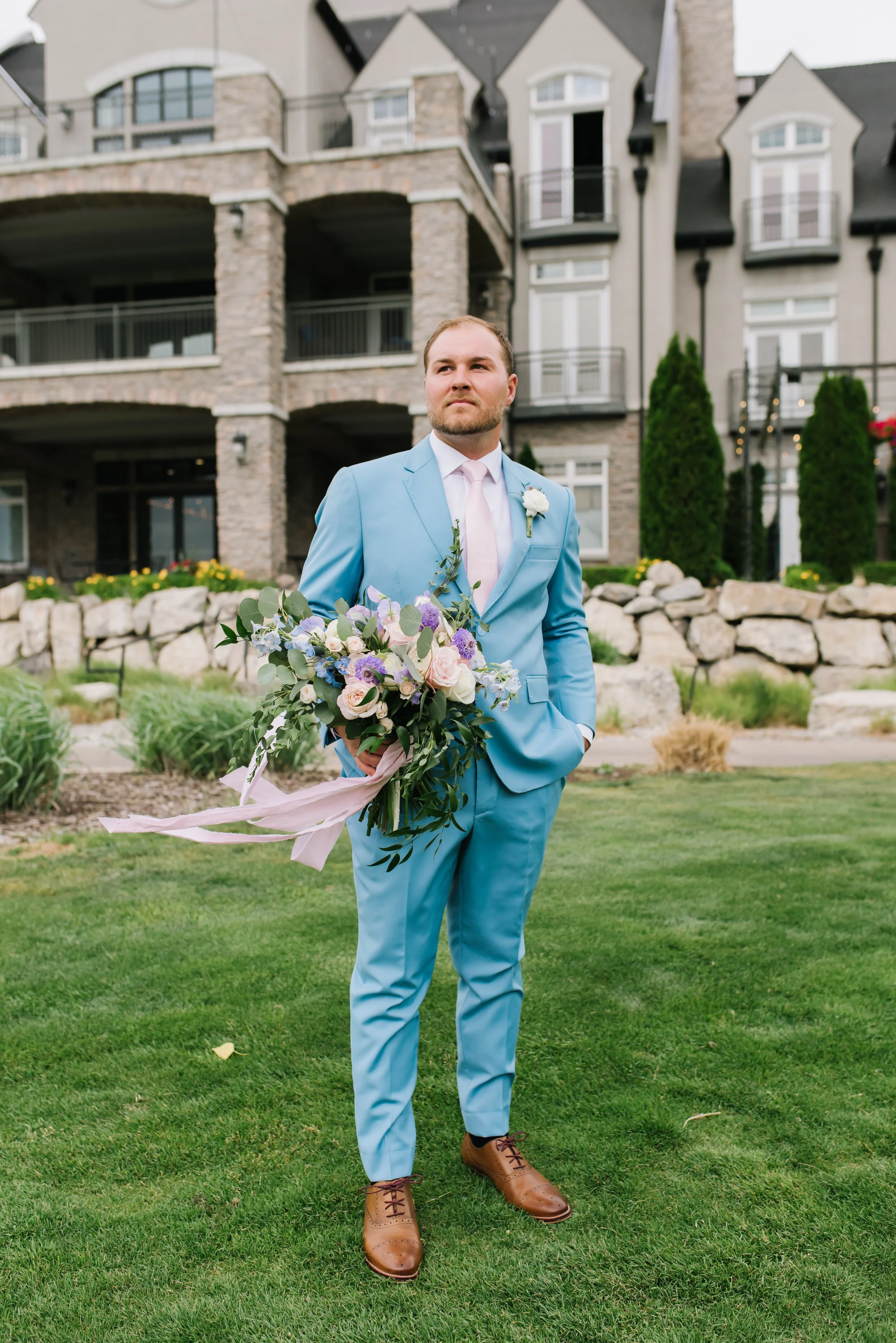 A man dressed in a light blue suit, pink tie, and brown shoes standing on a grassy lawn in front of a stone and stucco building. He is holding a bouquet of flowers with pink and purple blooms and green foliage.