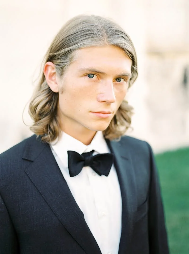 A young man with long, wavy silver hair and blue eyes wearing a black tuxedo with a white shirt and black bow tie.