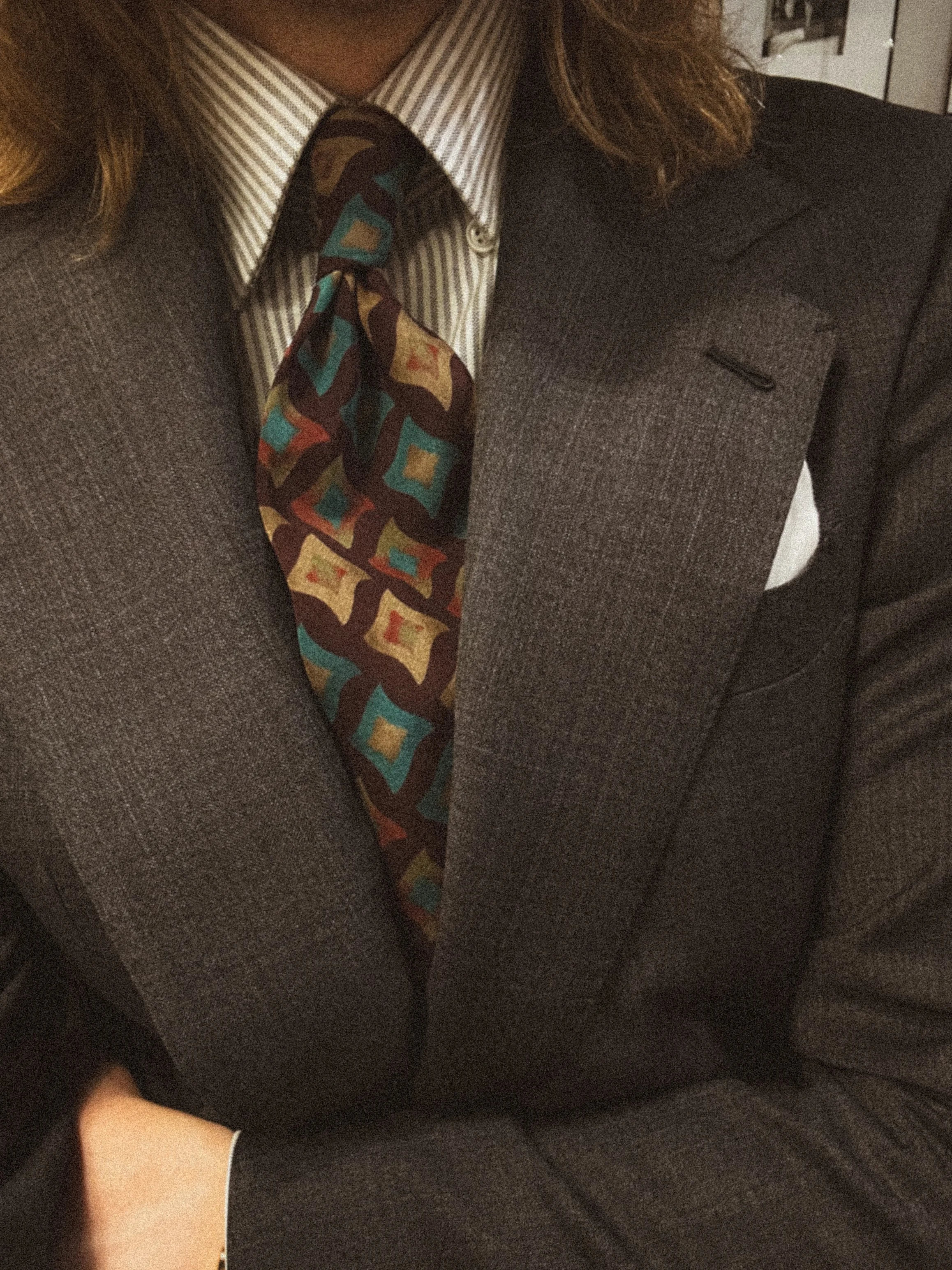 Close-up of a person wearing a charcoal suit, a patterned necktie, and a striped collared shirt, with long brown hair partially visible