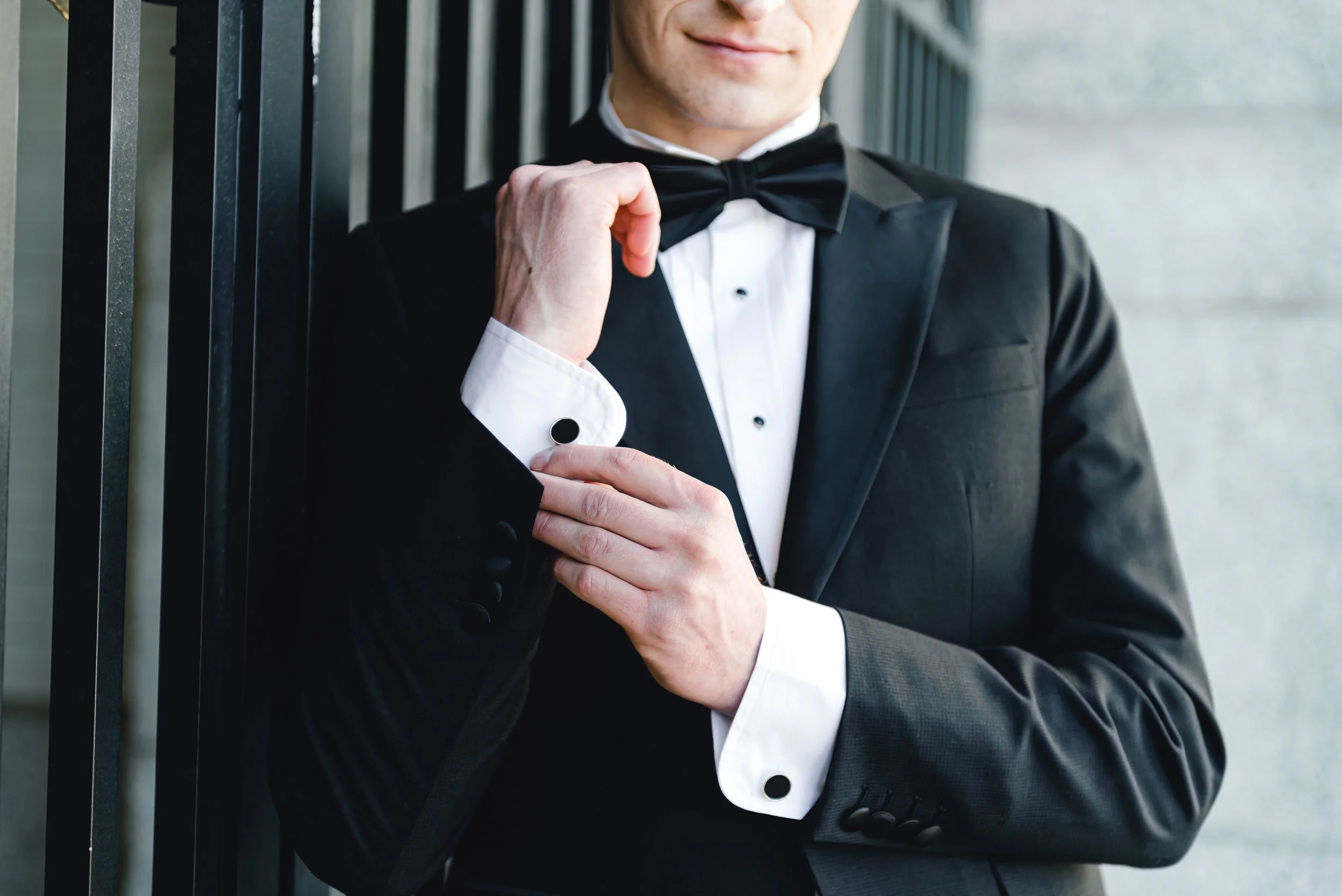 A man dressed in a tuxedo with a white shirt and black bow tie, adjusting his cufflink, standing near black metal bars.