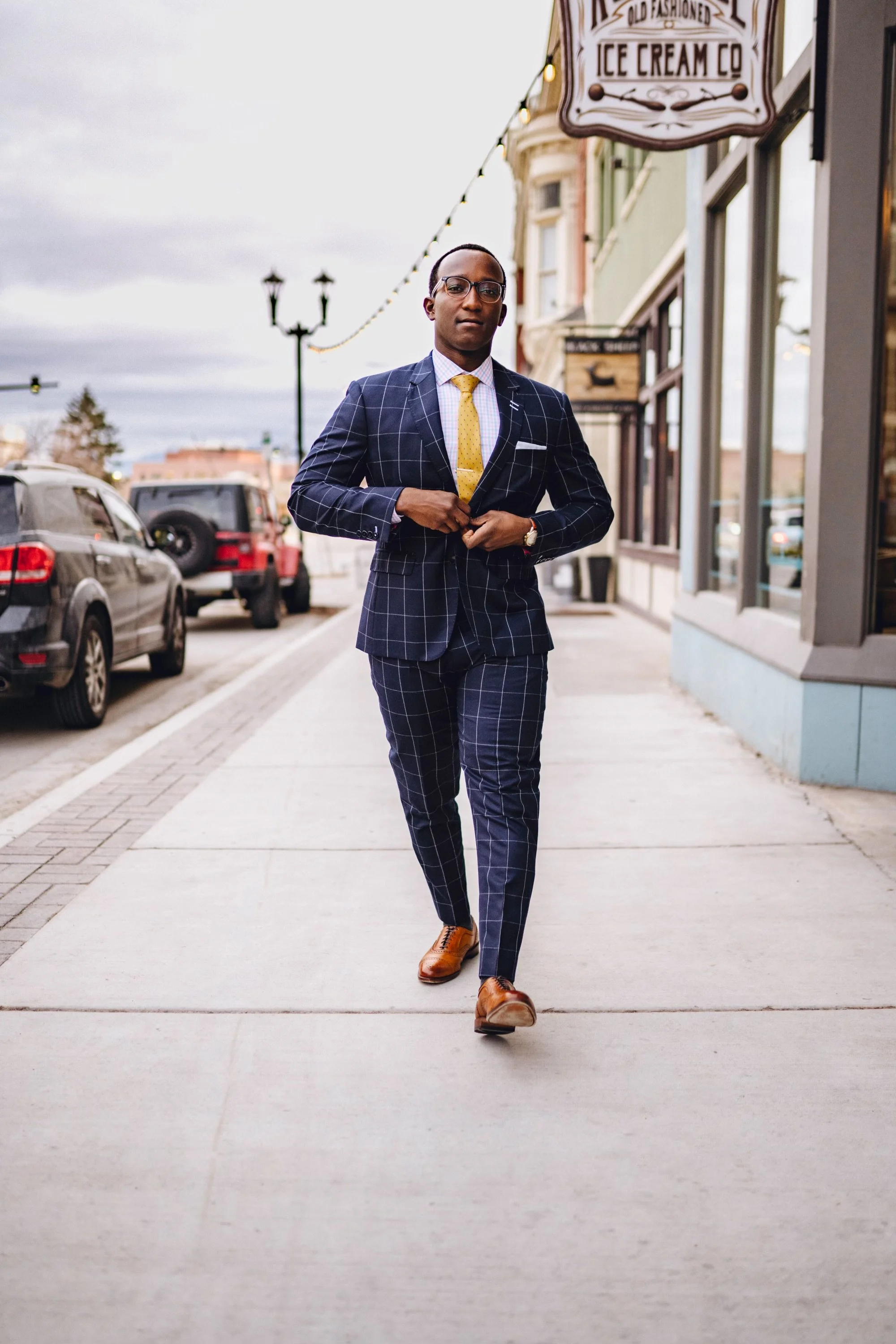 A man in a dark blue windowpane suit with a white shirt and yellow tie walking on a city sidewalk, adjusting his suit jacket. He is wearing brown shoes and glasses, with cars parked along the street and storefronts behind him.