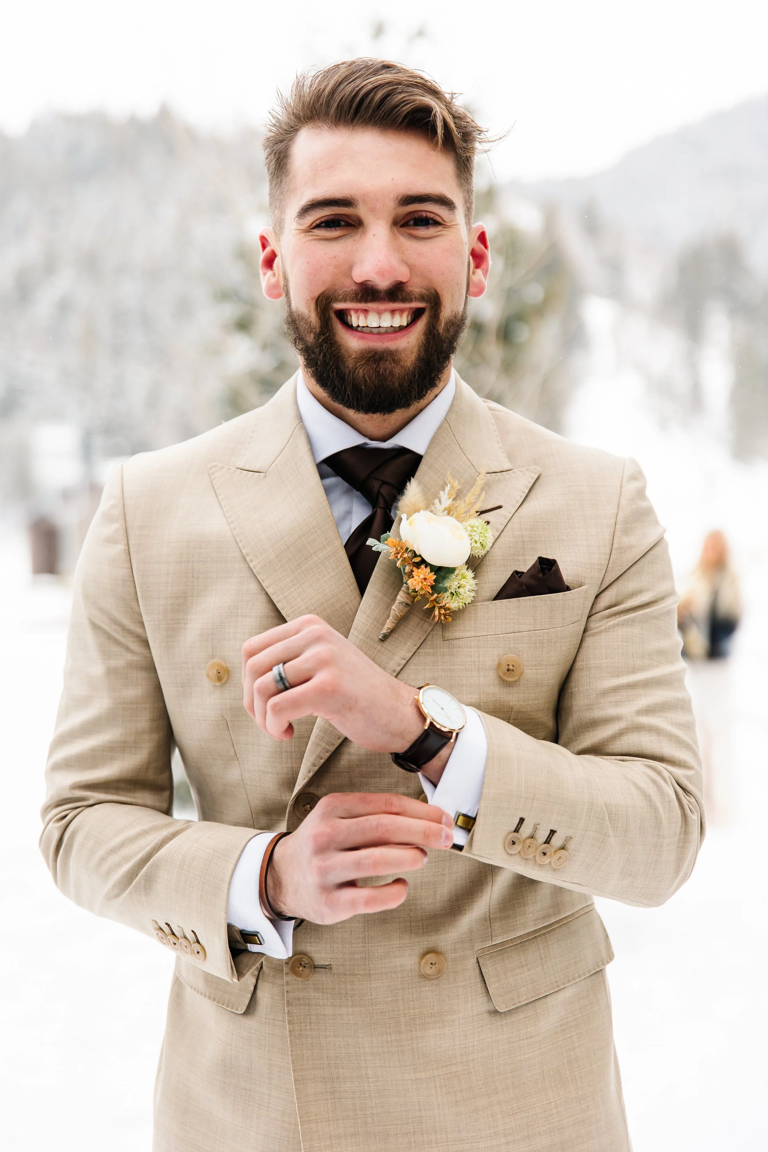 A smiling man dressed in a beige suit with a white shirt and dark tie, wearing a boutonniere and a watch, outdoors in winter with blurred snow-covered trees in the background.