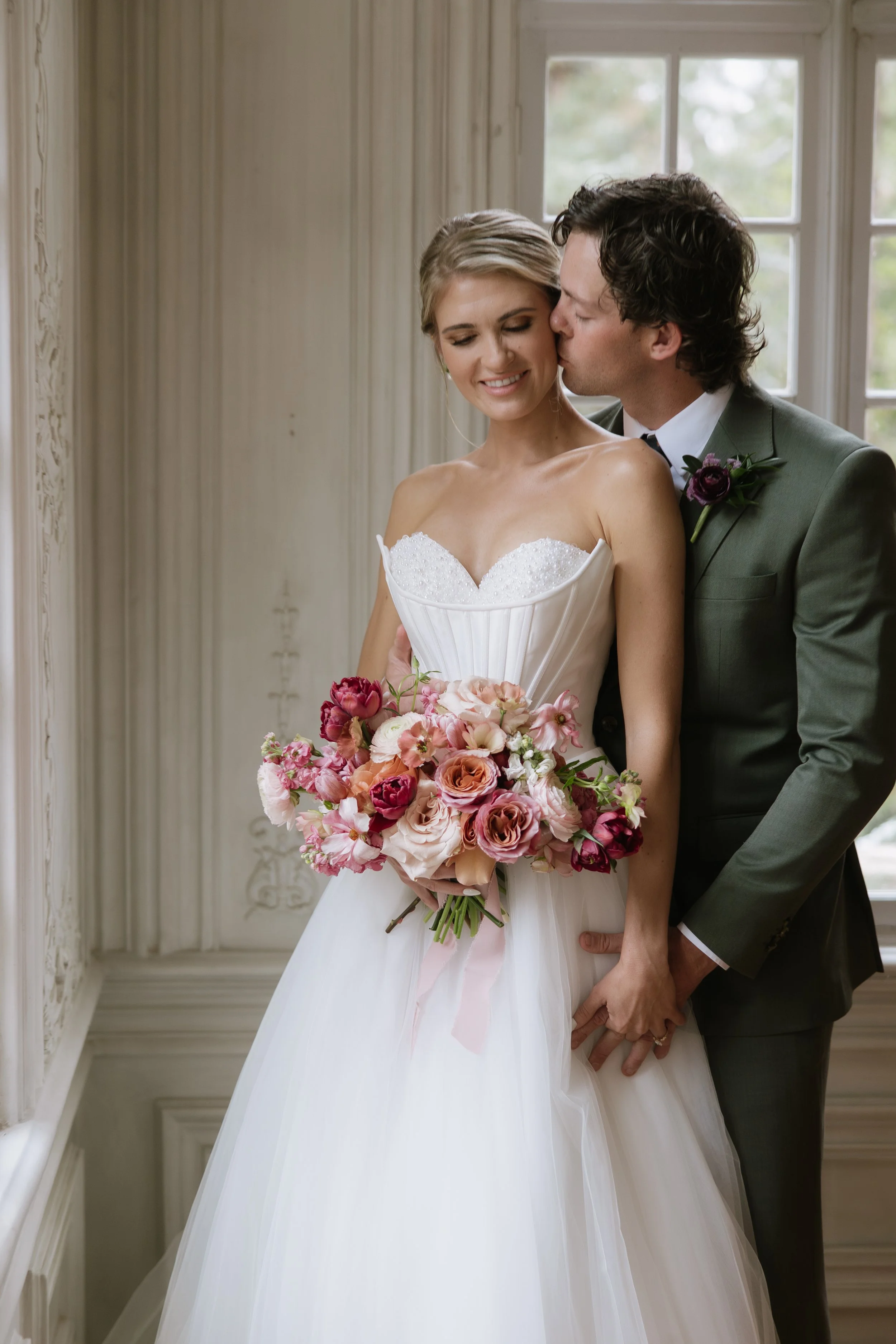 A bride and groom standing together in a room with large windows. The groom kisses the bride on the cheek, and she is holding a bouquet of pink, white, and purple flowers. The bride is wearing a strapless white wedding gown, and the groom is wearing 