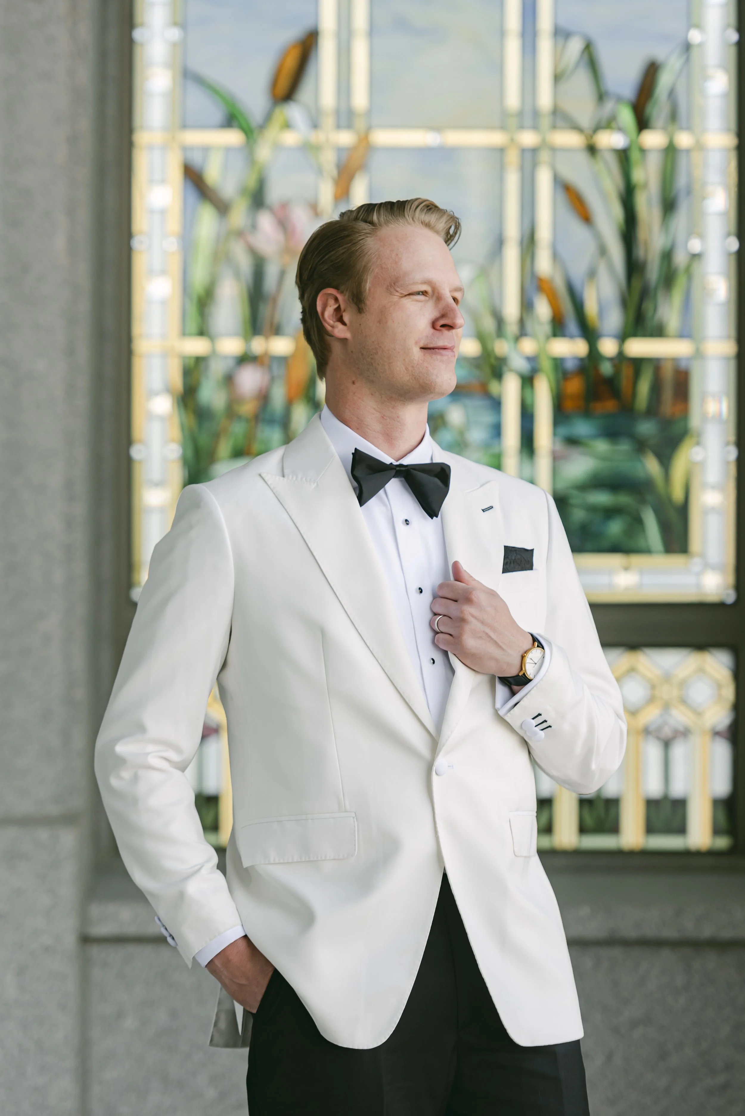 A man wearing a white tuxedo and black bow tie, standing indoors with a colorful stained glass window and greenery in the background.