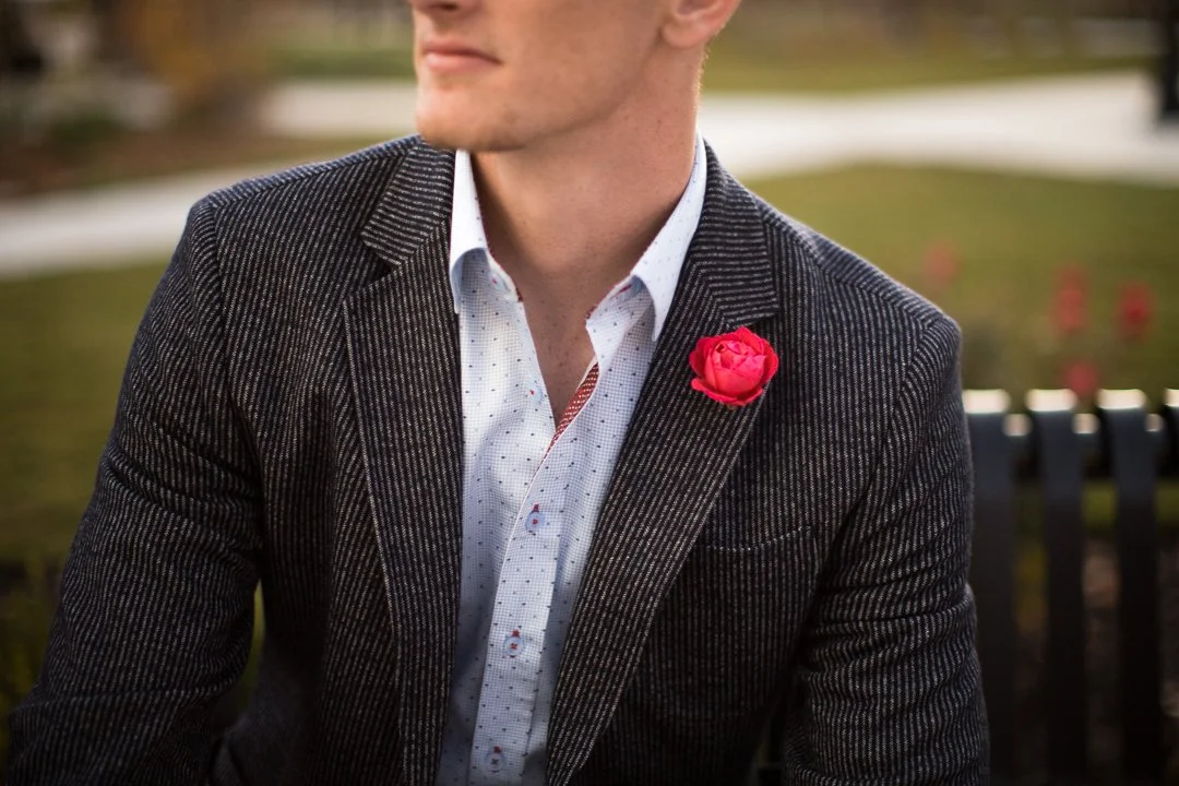A man wearing a dark, pinstriped suit and a white shirt with small red and blue patterns, sitting outdoors in a park with a red rose boutonnière on his blazer.