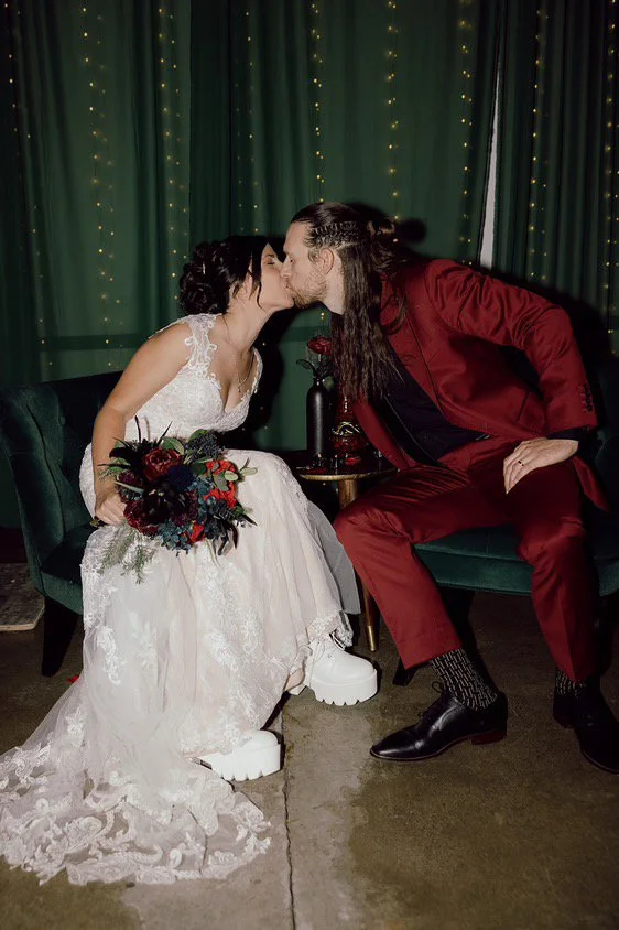 A wedding couple sharing a kiss inside, the bride sitting on a green chair with a bouquet, the groom in a red suit sitting on a matching chair, with green curtains and string lights in the background.