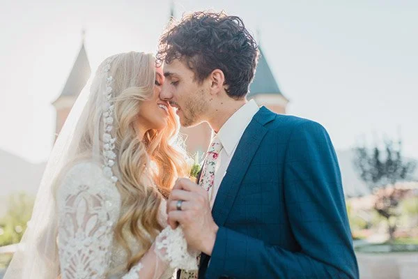 A newlywed couple with their foreheads touching outdoors during daytime, with a castle-like building in the background.