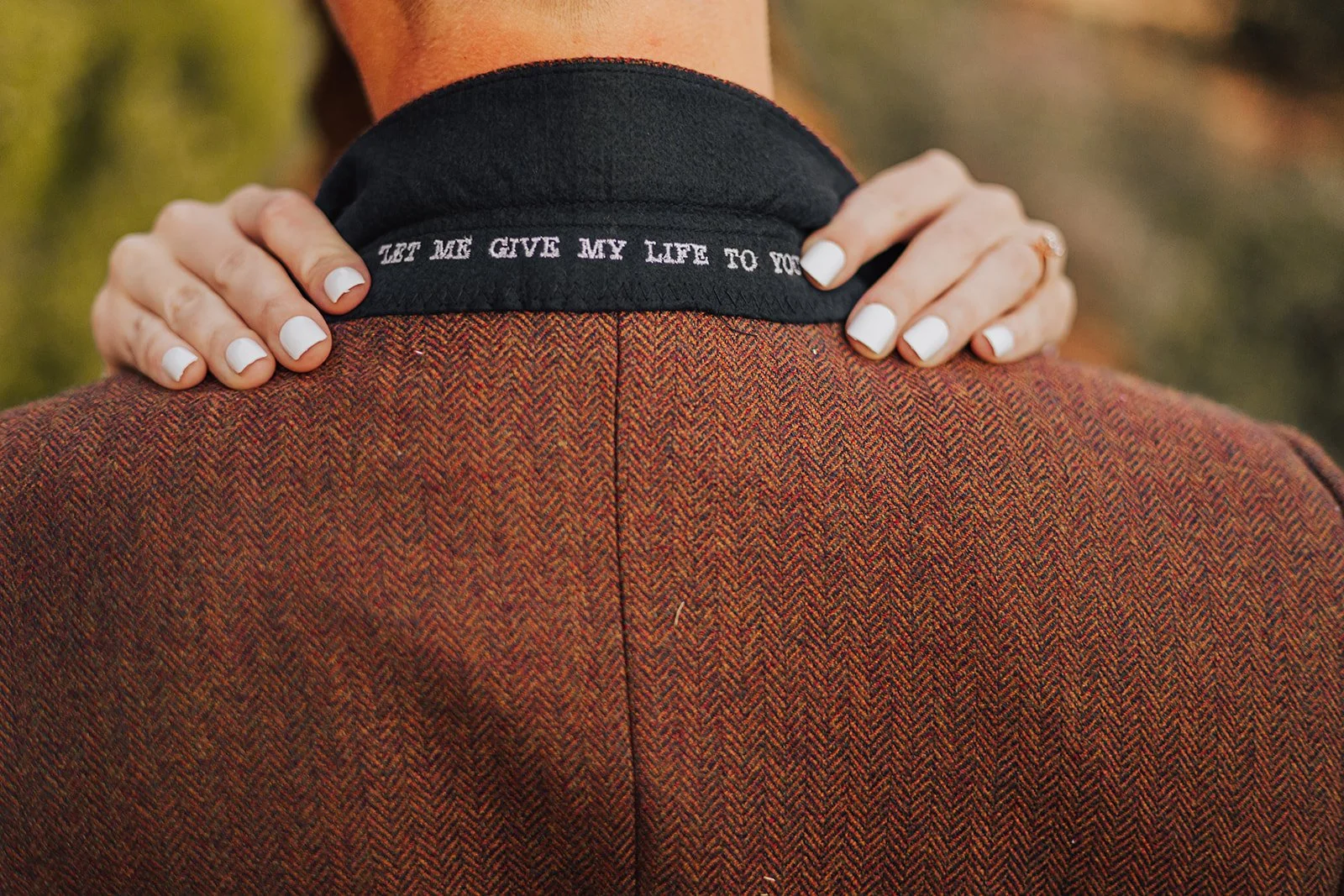 Close-up of a person's hands resting on their shoulders, wearing a herringbone jacket with black under collar lining with white embroidered text: 'Let me give my life to you'