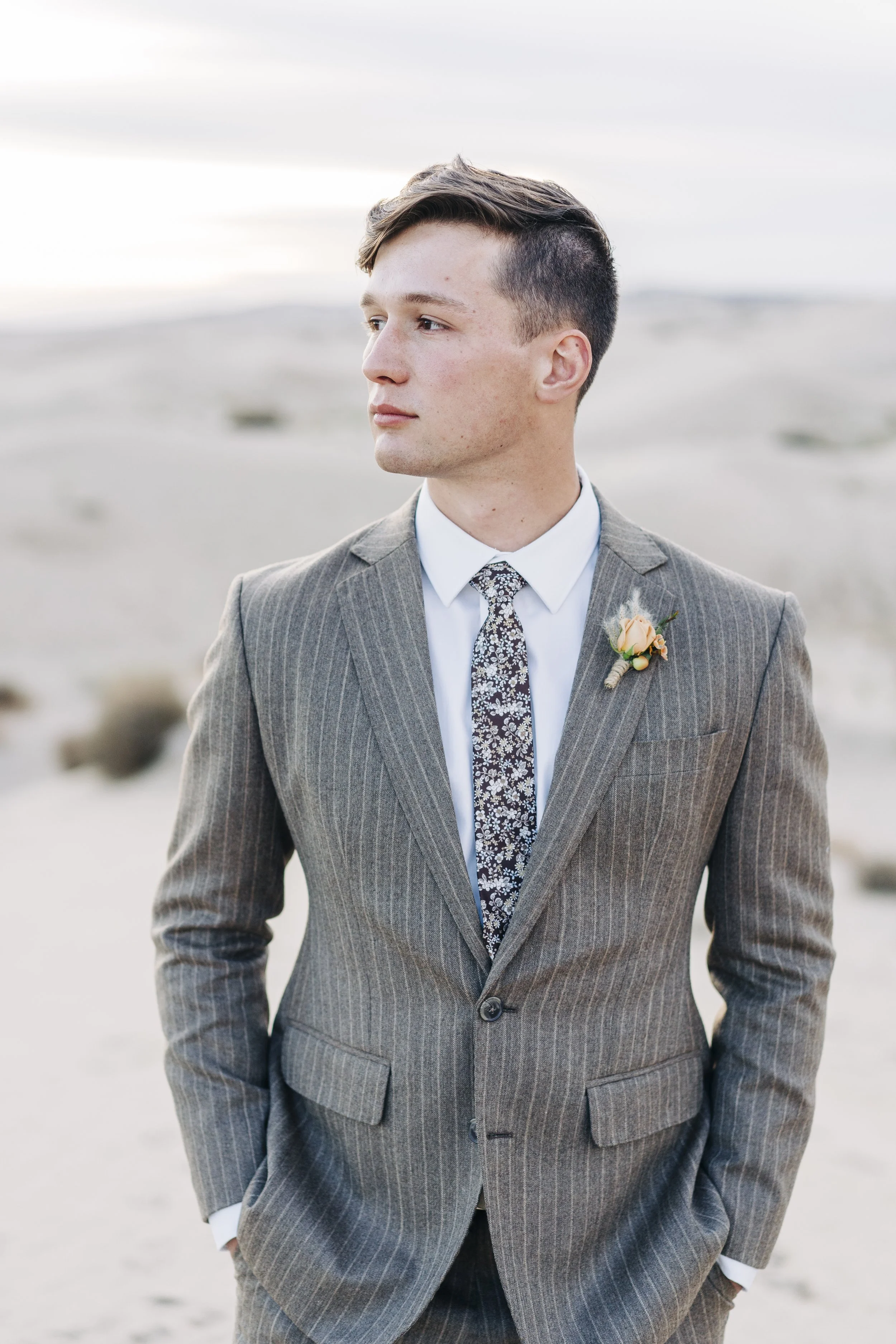 A young man in a gray pinstripe suit, white shirt, and patterned tie, with a boutonniere on his lapel, standing outdoors on a beach with a neutral expression as he looks to the side.