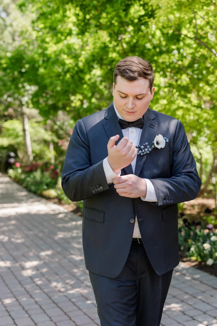 A man in a navy suit adjusting his cufflink outdoors with green trees and a brick pathway in the background.