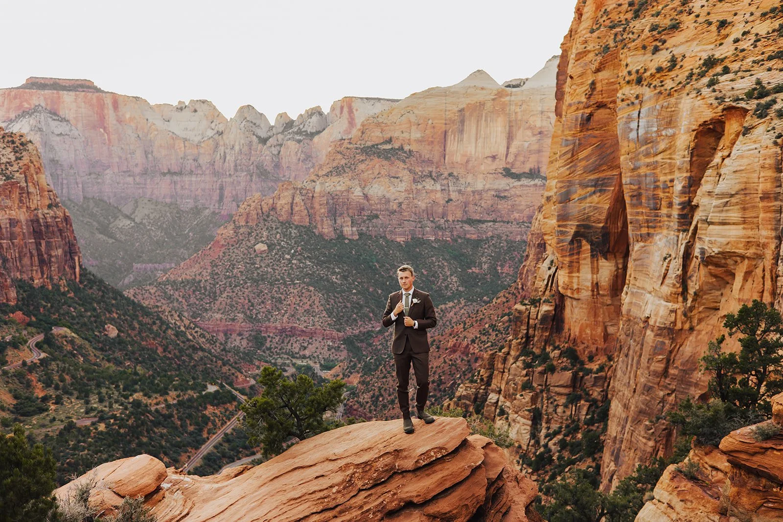 A man in a dark suit and tie stands on a rocky ledge in a canyon, past the towering colorful cliffs and mountains of Zion National Park.