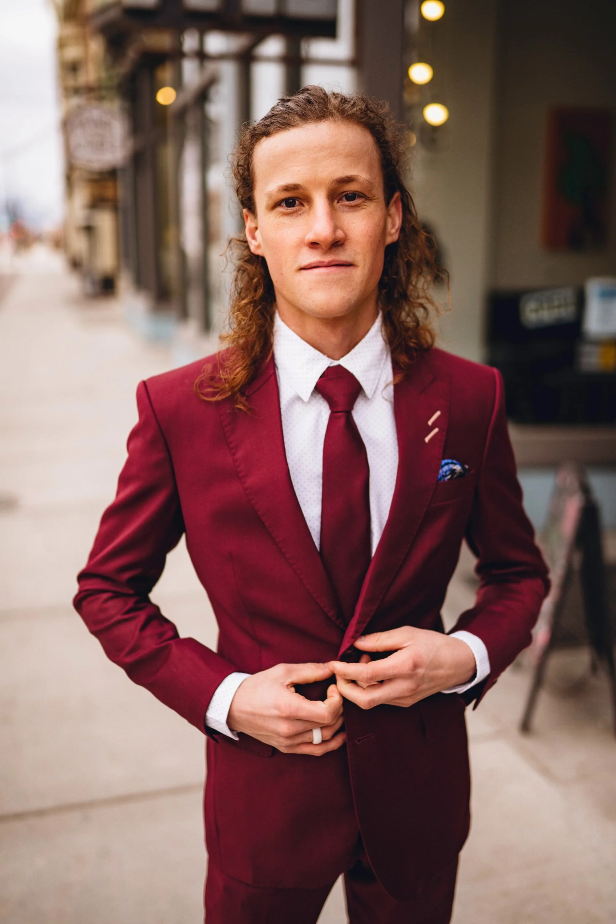A young man in a burgundy suit, white shirt, and burgundy tie standing outdoors, adjusting his suit jacket.
