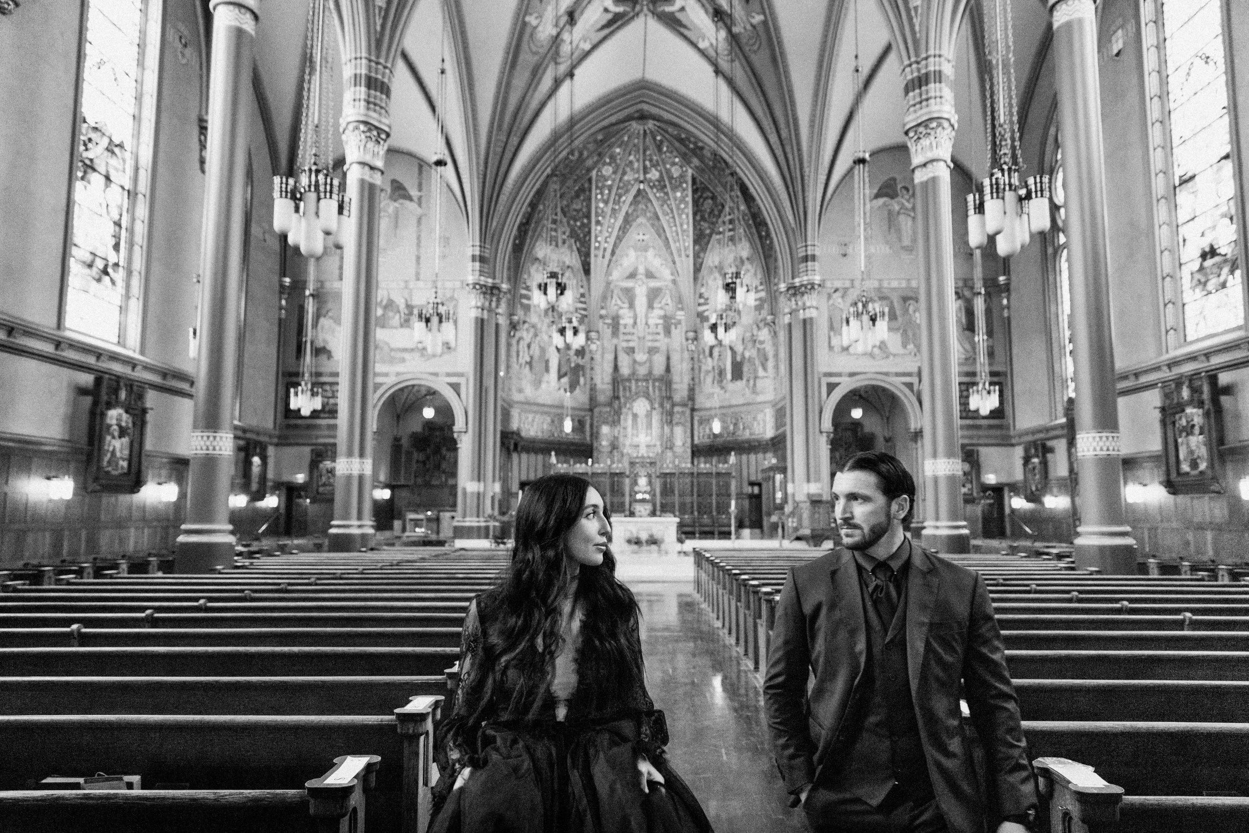 A church interior with high vaulted ceilings, ornate chandeliers, and stained glass windows. A woman with long dark hair and a man with short dark hair and a beard are walking down the aisle for their wedding.