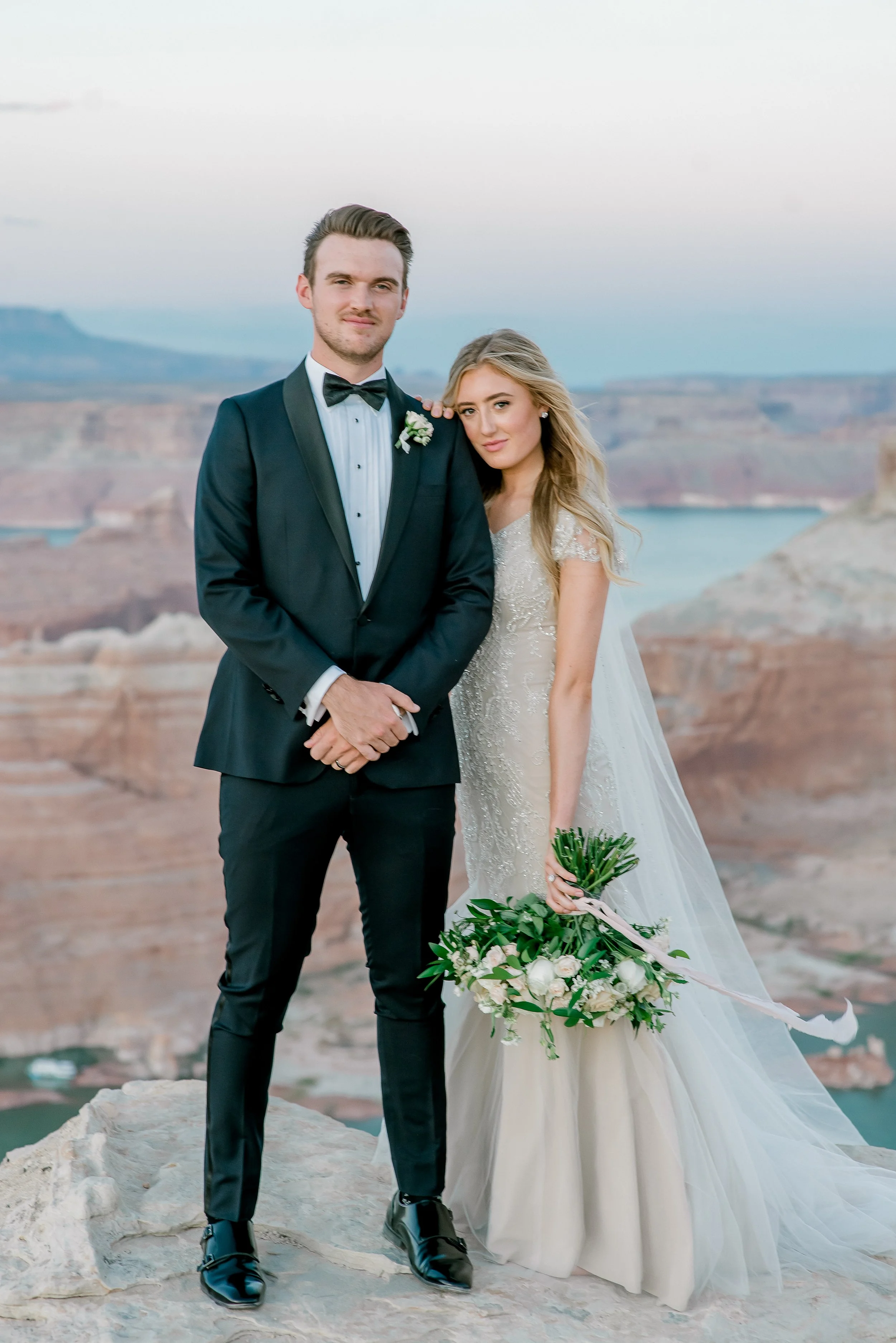 A bride and groom standing together on a rocky landscape with a canyon and a body of water in the background during sunset. The groom is wearing a tuxedo with a bowtie, and the bride is in a wedding gown holding a bouquet of flowers.