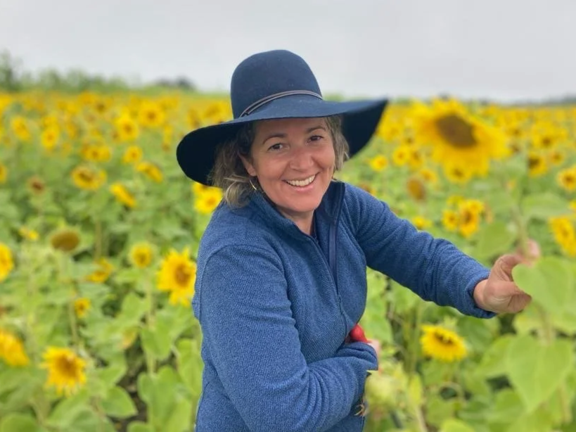 Woman smiling in a sunflower field, wearing a wide-brimmed hat and blue jacket.