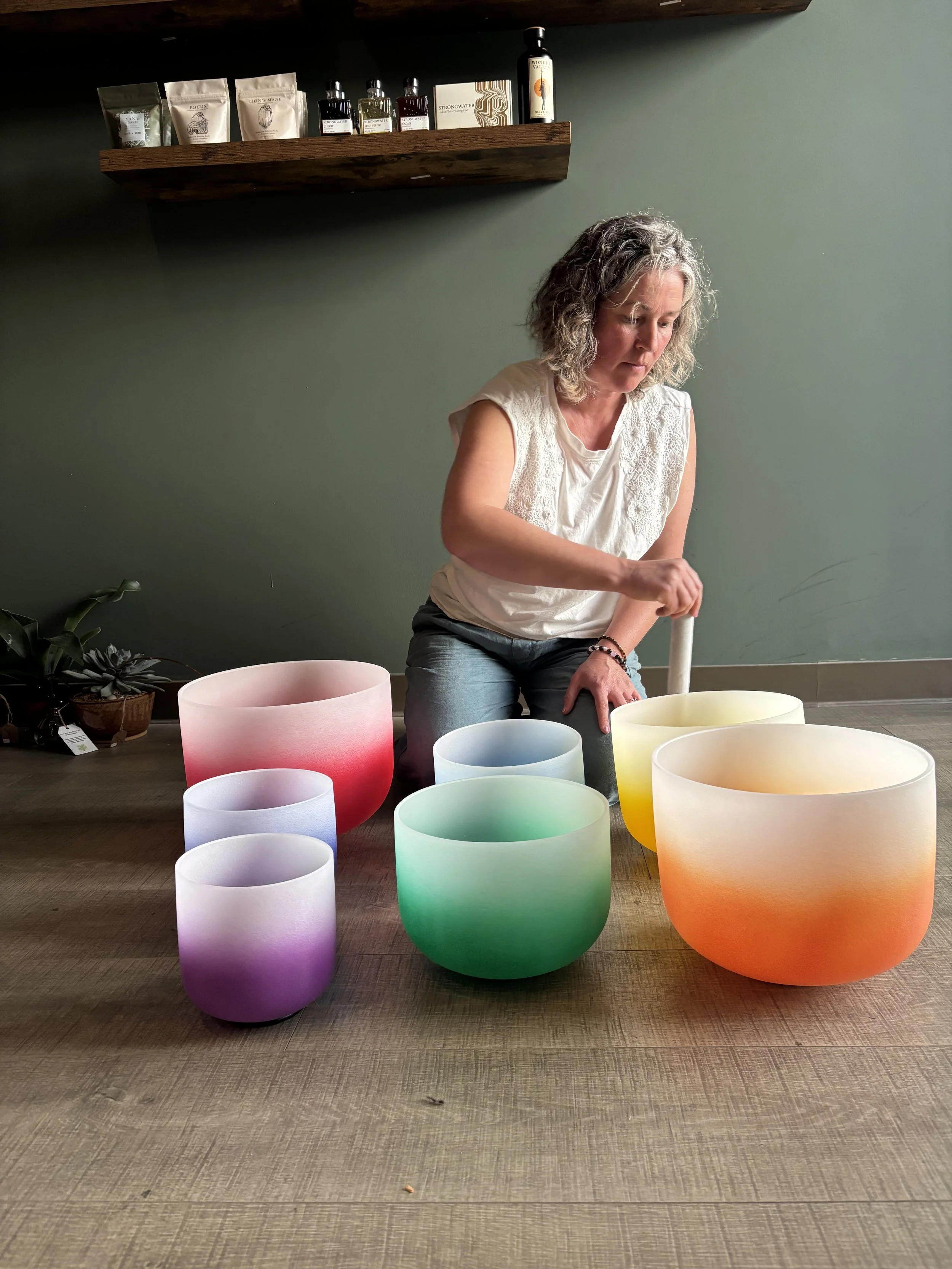 A woman with curly gray hair playing a crystal singing bowl, surrounded by colorful illuminated bowls on a wooden floor in a room with a green wall and wooden shelf with bottles and packages.