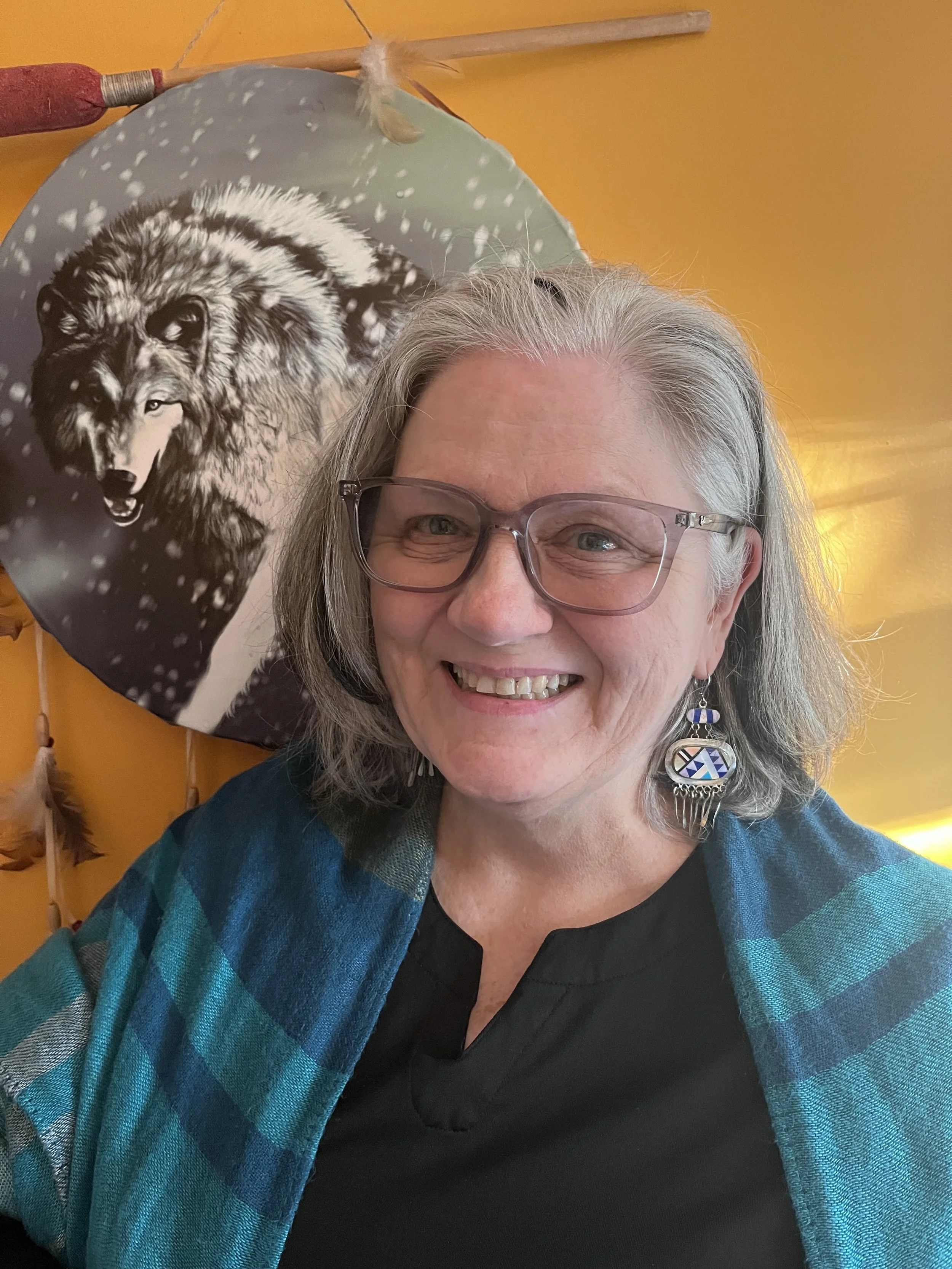 A woman with glasses smiling in front of a colorful wall with a wolf-themed balloon. She is wearing a blue and black plaid shawl over a black top and large earrings.
