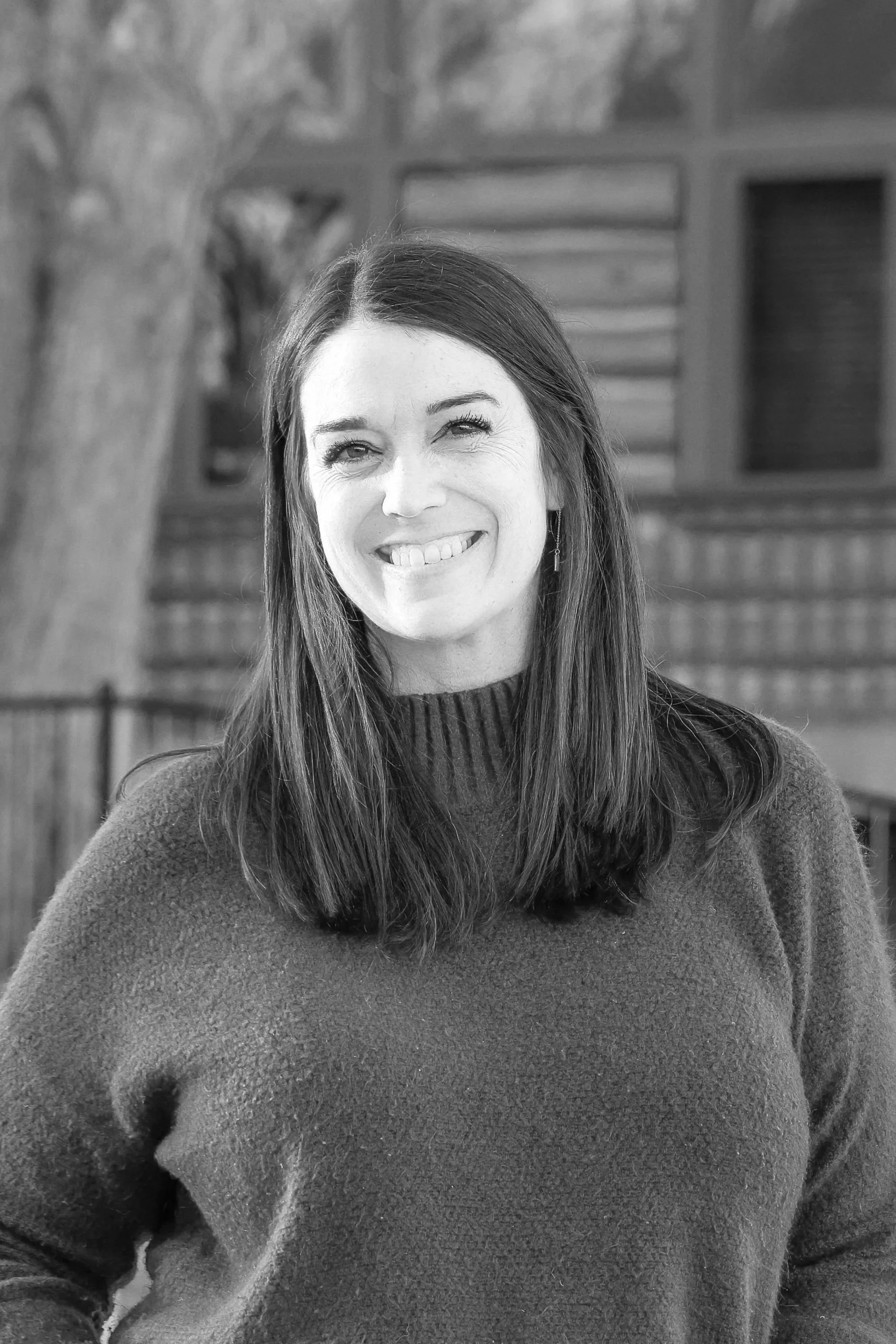 Black and white photo of a woman with long, straight hair, smiling with teeth showing, wearing a turtleneck sweater, in front of a wooden house with window shutters and a tree in the background.